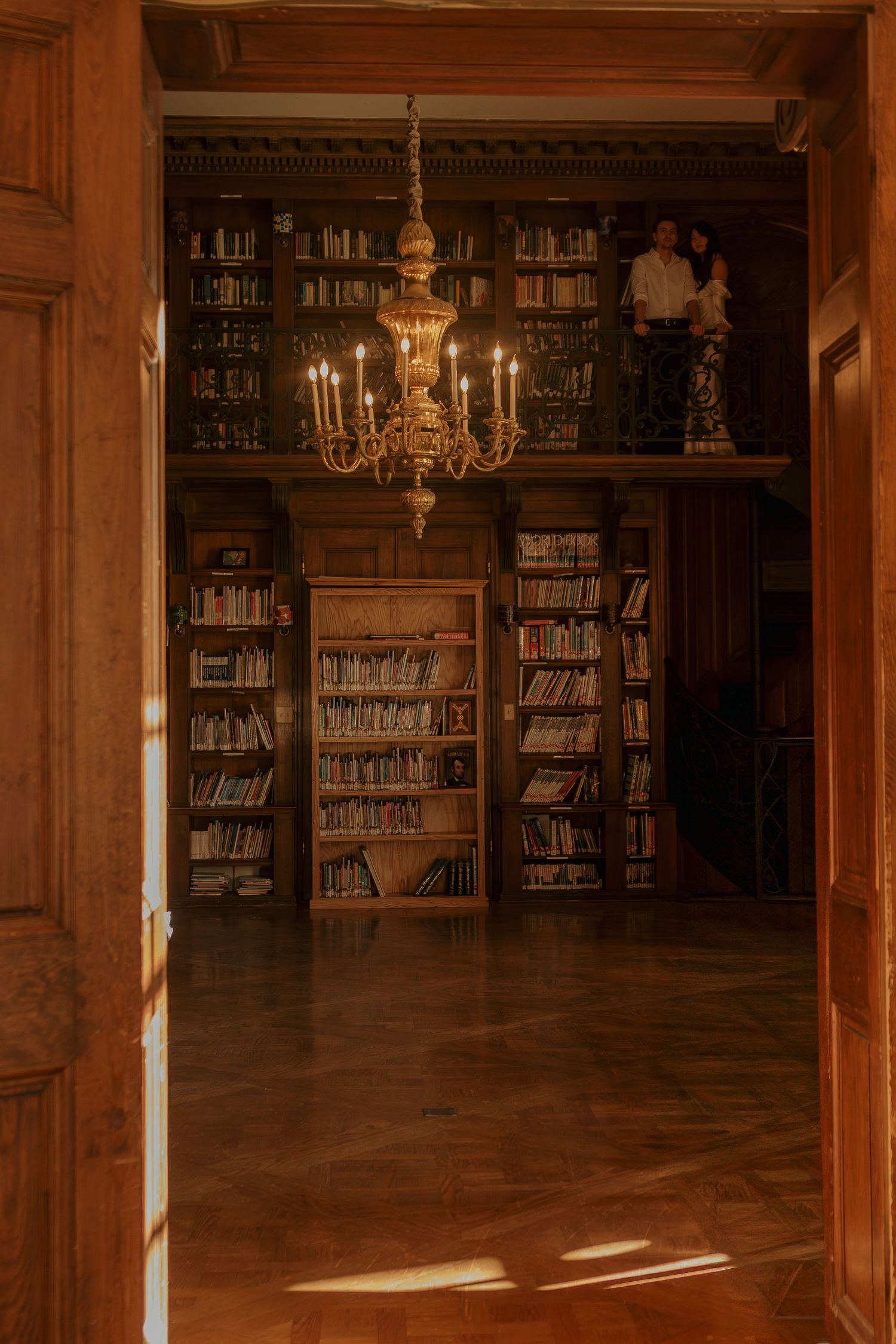 Elegant library interior with wooden bookshelves and ornate brass chandelier casting warm light across hardwood floors.