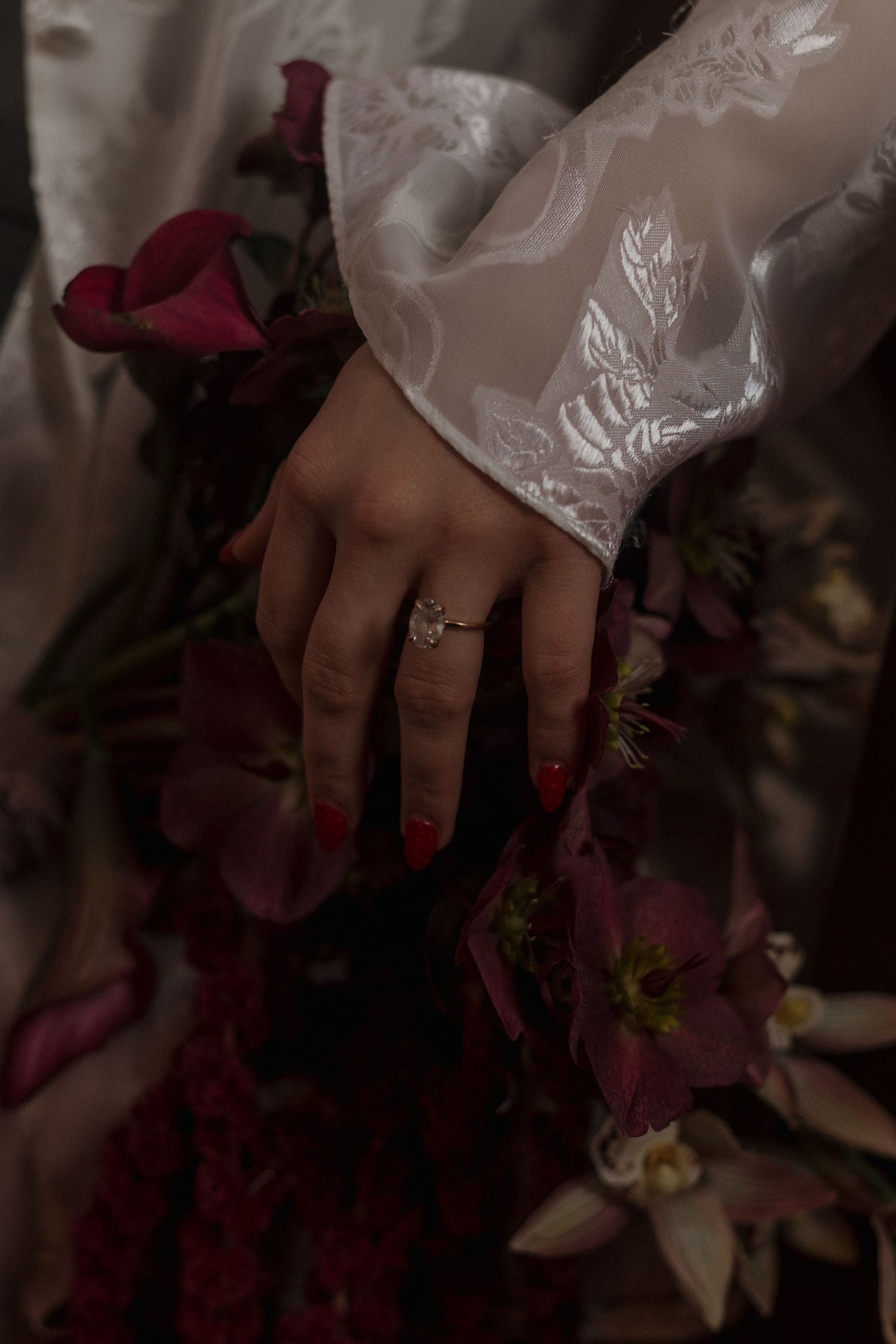 A delicate lace sleeve and diamond ring against deep red flowers in moody lighting.