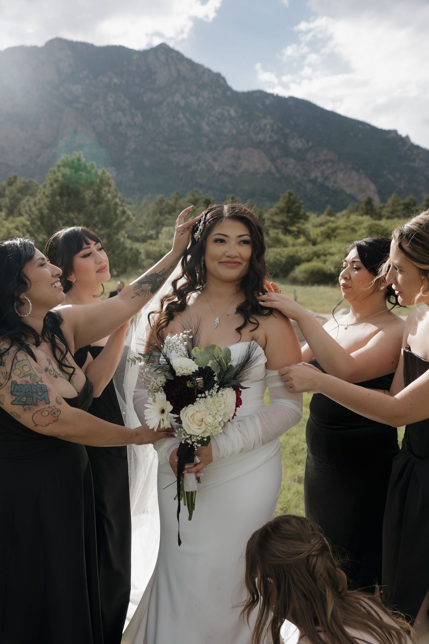 Bride in white gown holds bouquet as bridesmaids in black dresses help her prepare with mountains behind.