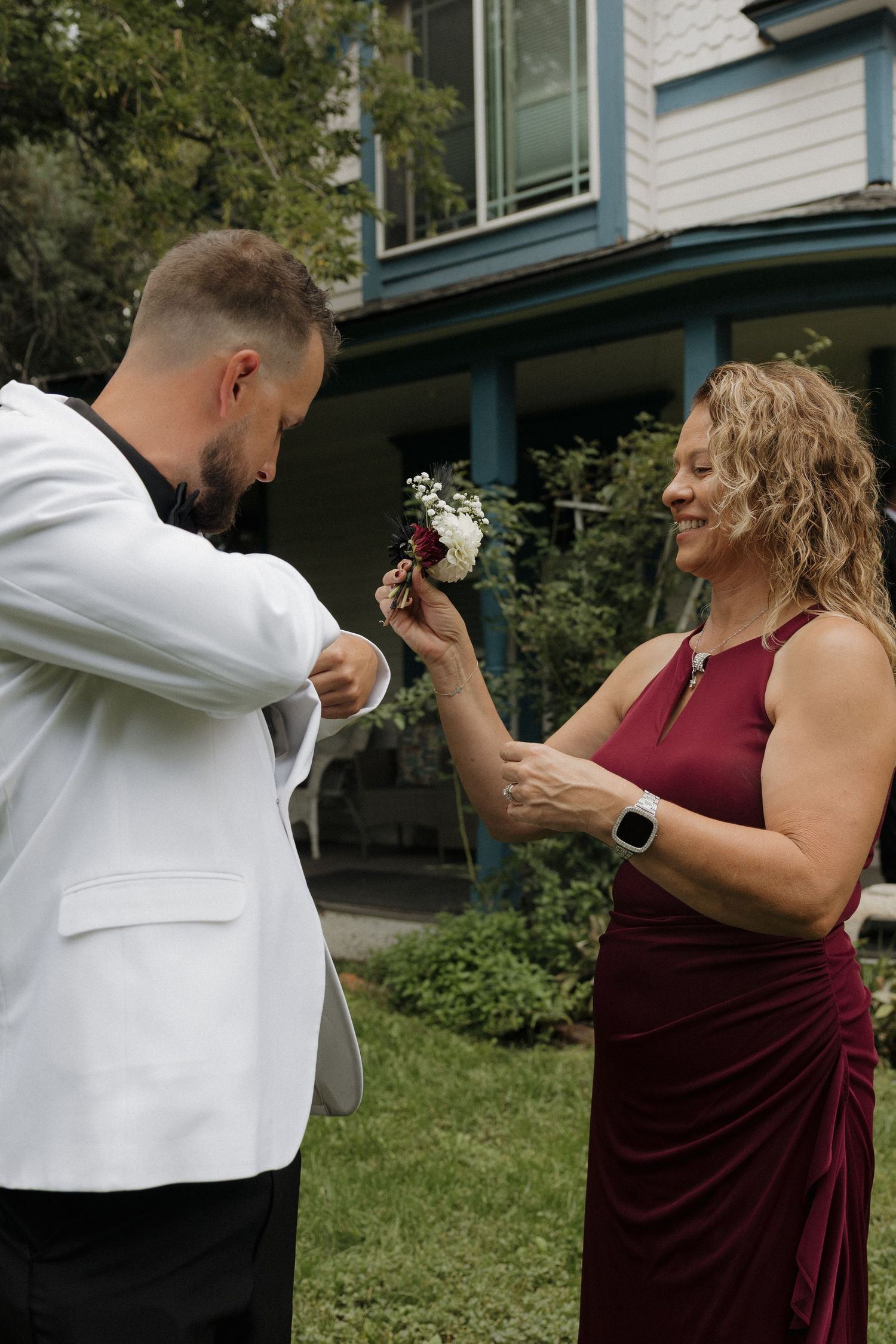 Woman in burgundy dress pins boutonniere on man in white tuxedo jacket outdoors before wedding ceremony.