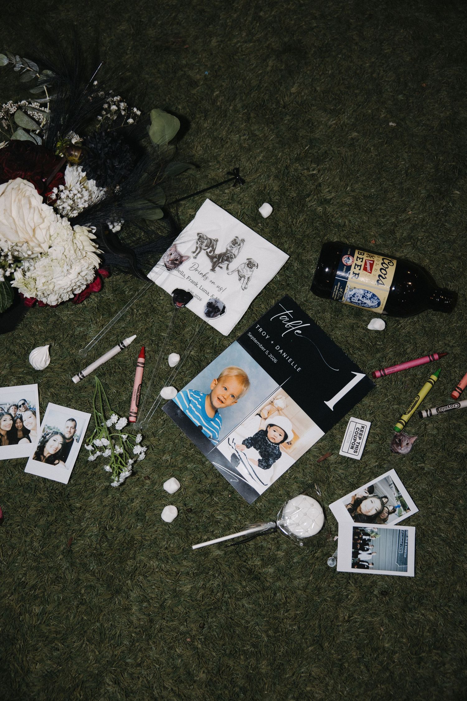 Flat lay of wedding mementos including Polaroids, flowers, crayons, and program on grass.