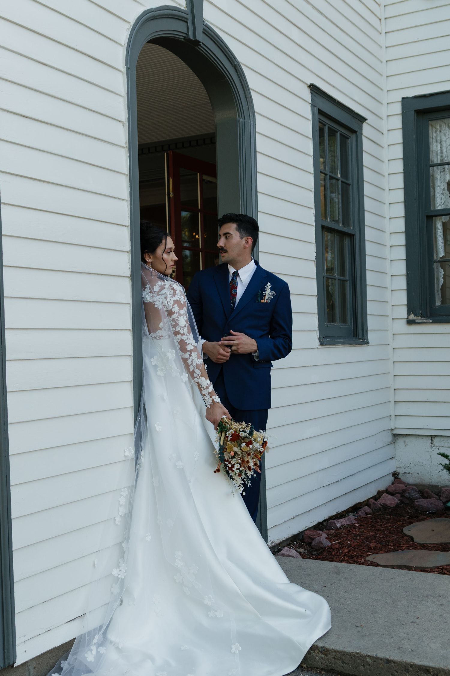 Wedding couple poses against white building with arched doorways during their first look.