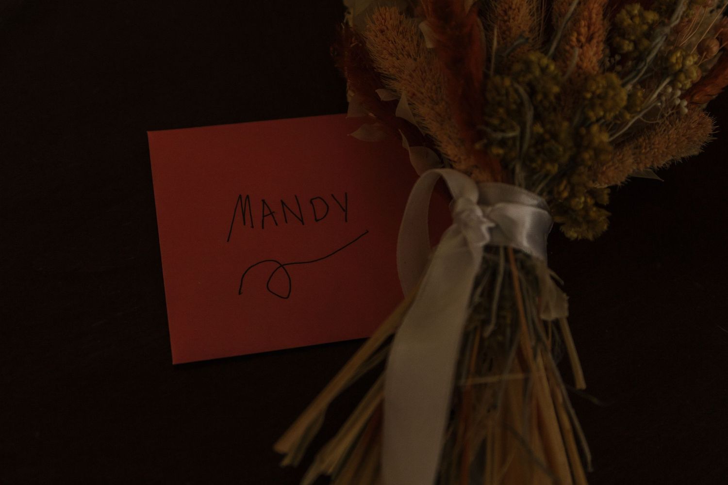 A red place card tied to a dried floral bouquet with a white ribbon in dark lighting.
