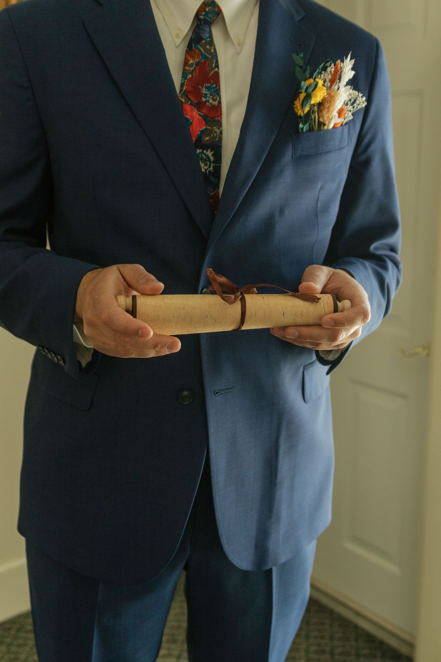 Close-up of groom in navy suit holding a rolled document with a colorful floral boutonniere.