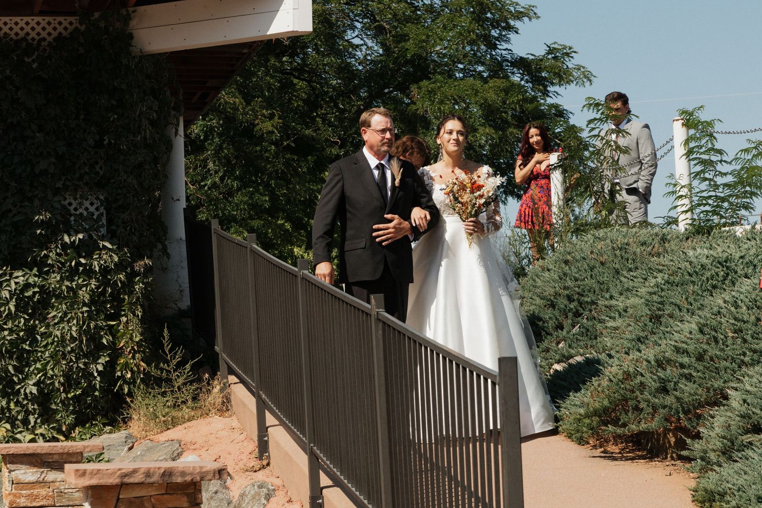 A wedding processional descends a ramp lined with black railings on a sunny day.