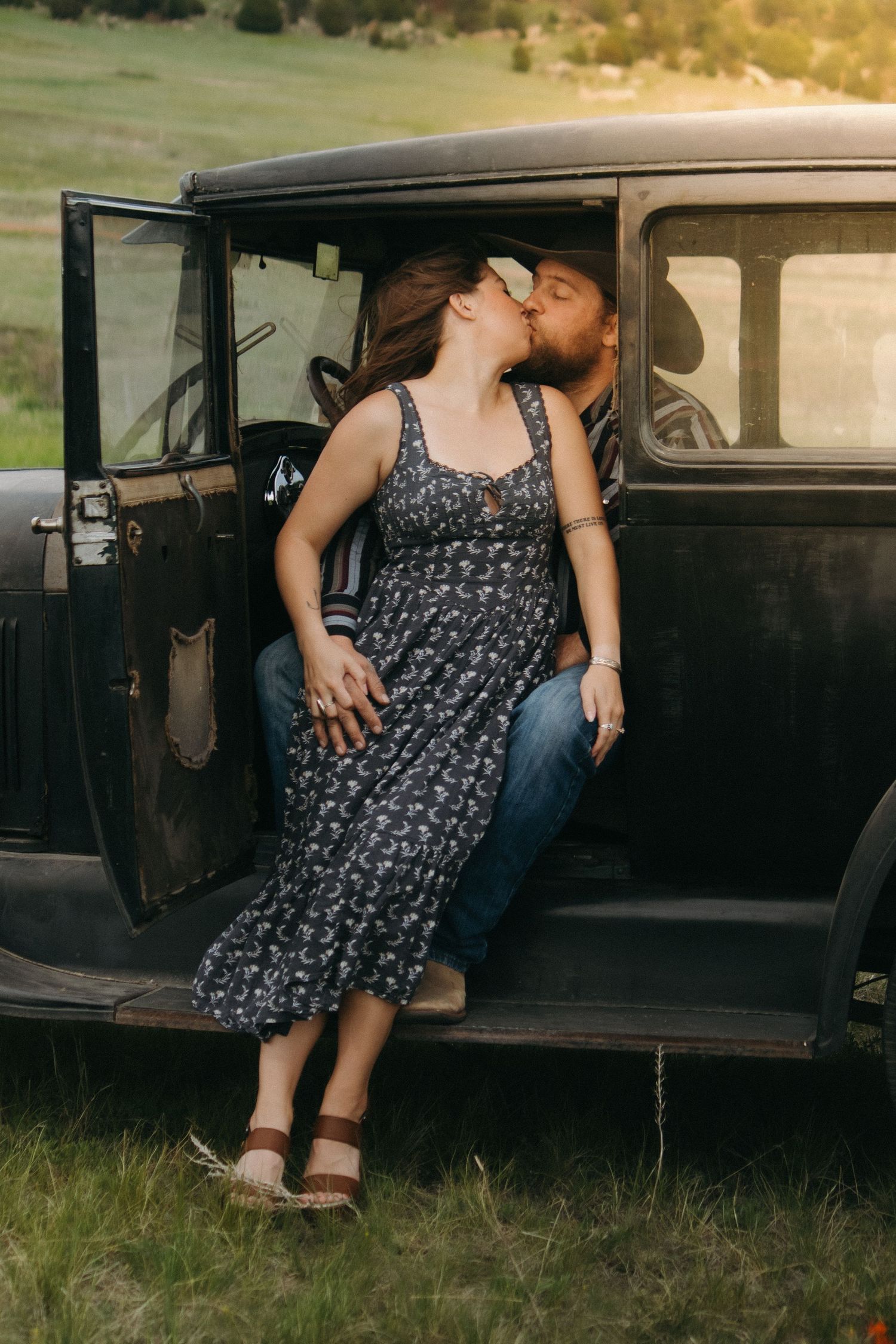 A couple sharing a romantic kiss while sitting in a vintage black car at sunset.