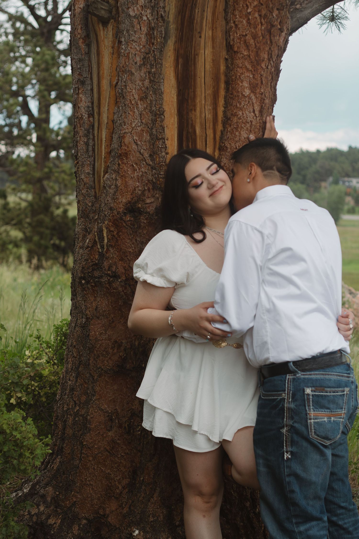 Intimate moment captured between couple against textured tree trunk in mountain landscape.