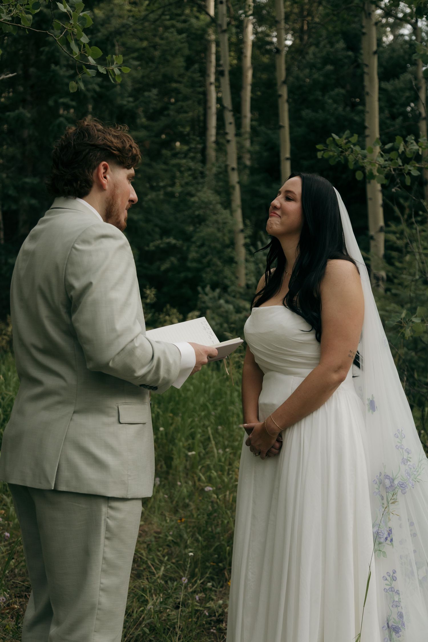 Groom reads heartfelt vows from a book to his smiling bride in a lush outdoor forest ceremony.