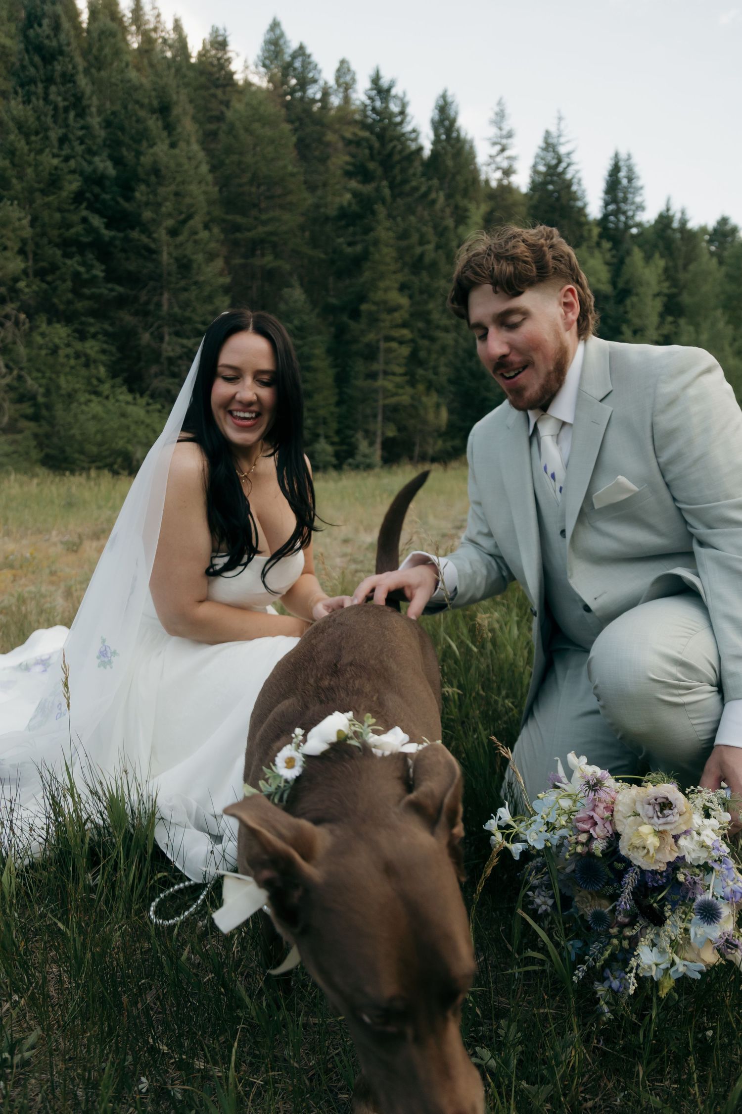 Smiling bride and groom pet their brown dog wearing a floral crown during outdoor wedding.