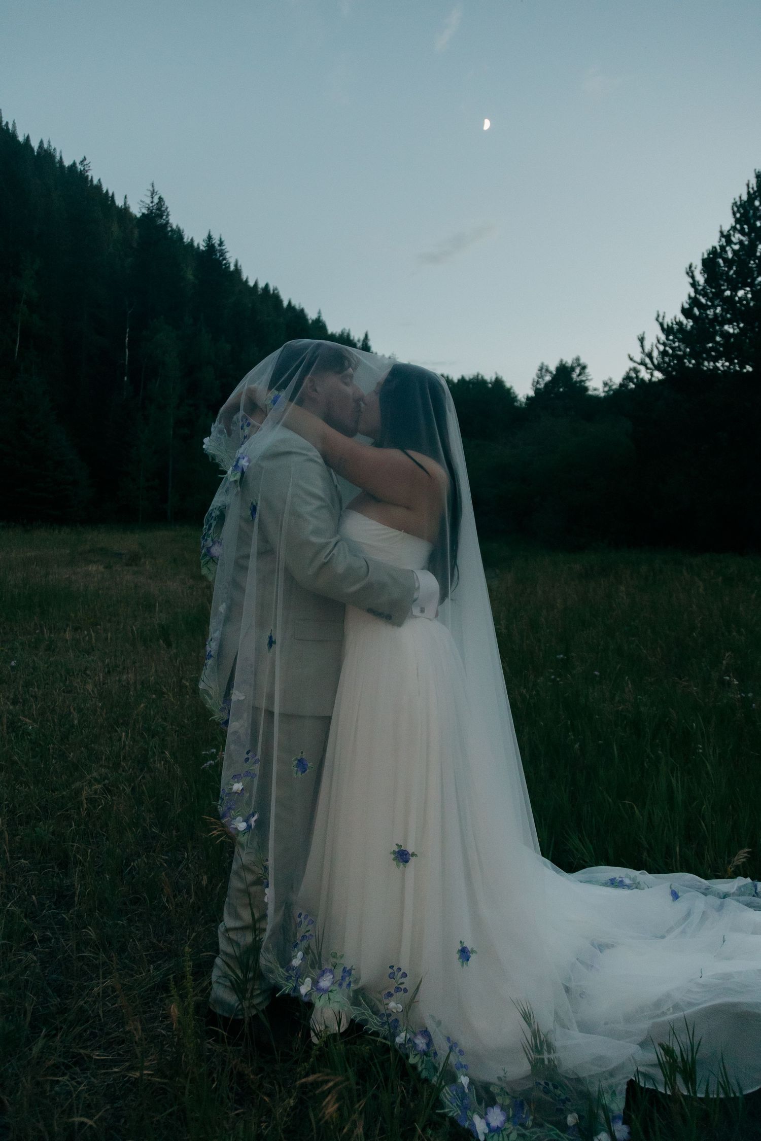 Bride and groom sharing a kiss under a floral veil in a meadow with forest backdrop at twilight.