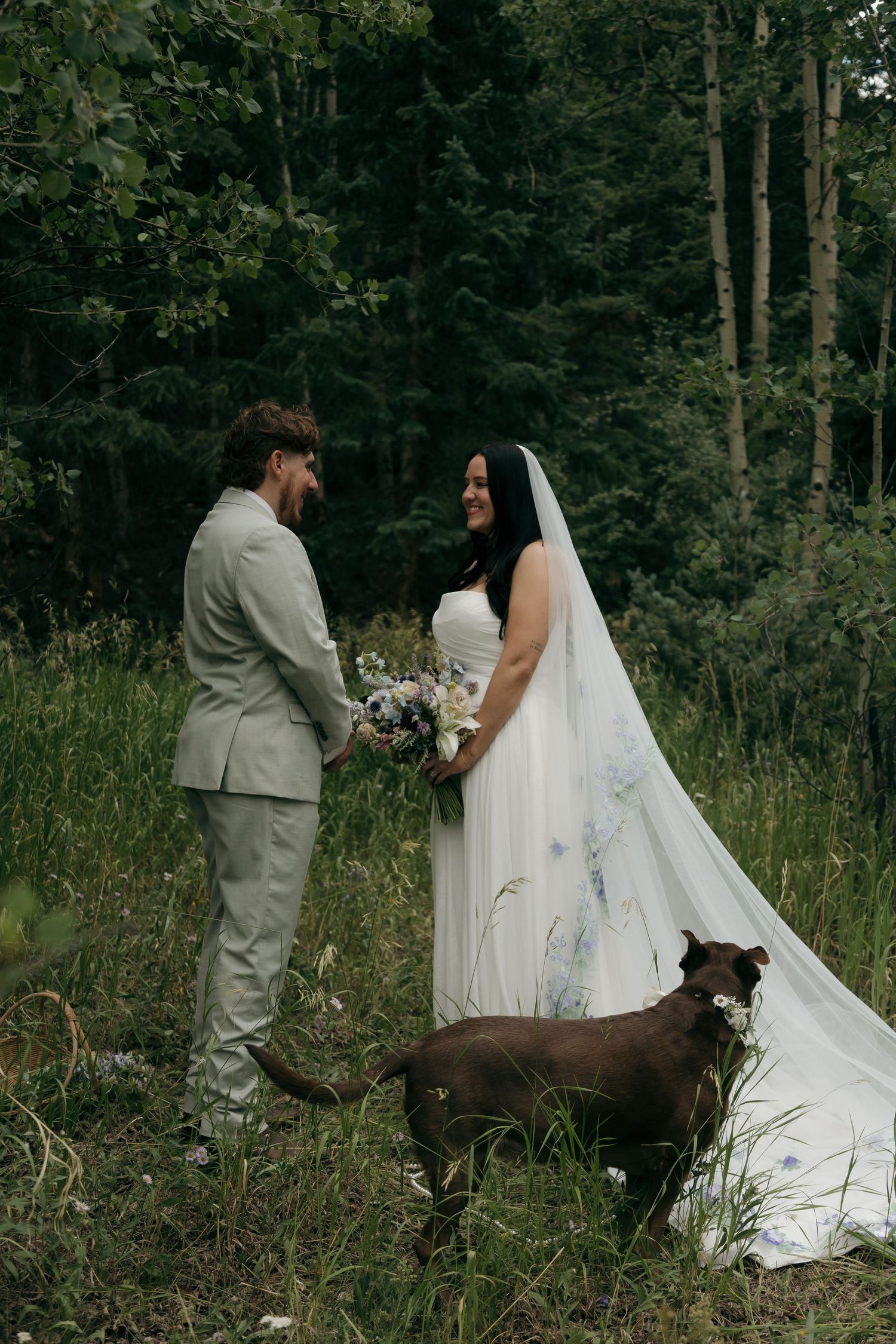 Smiling bride and groom stand together in a woodland setting with their chocolate Labrador dog nearby.
