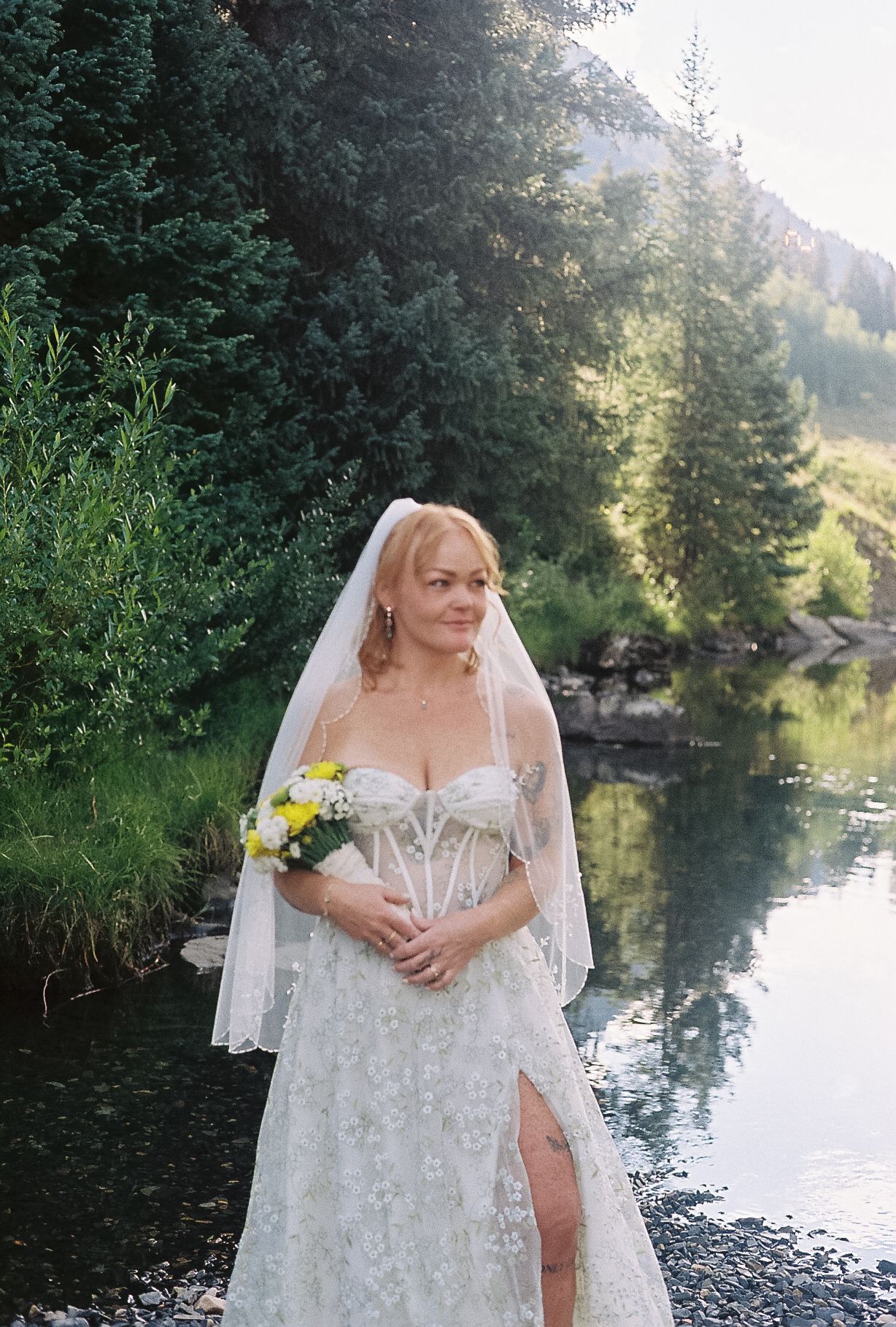 Radiant bride in lace corset wedding gown holding yellow bouquet beside a reflective mountain lake.