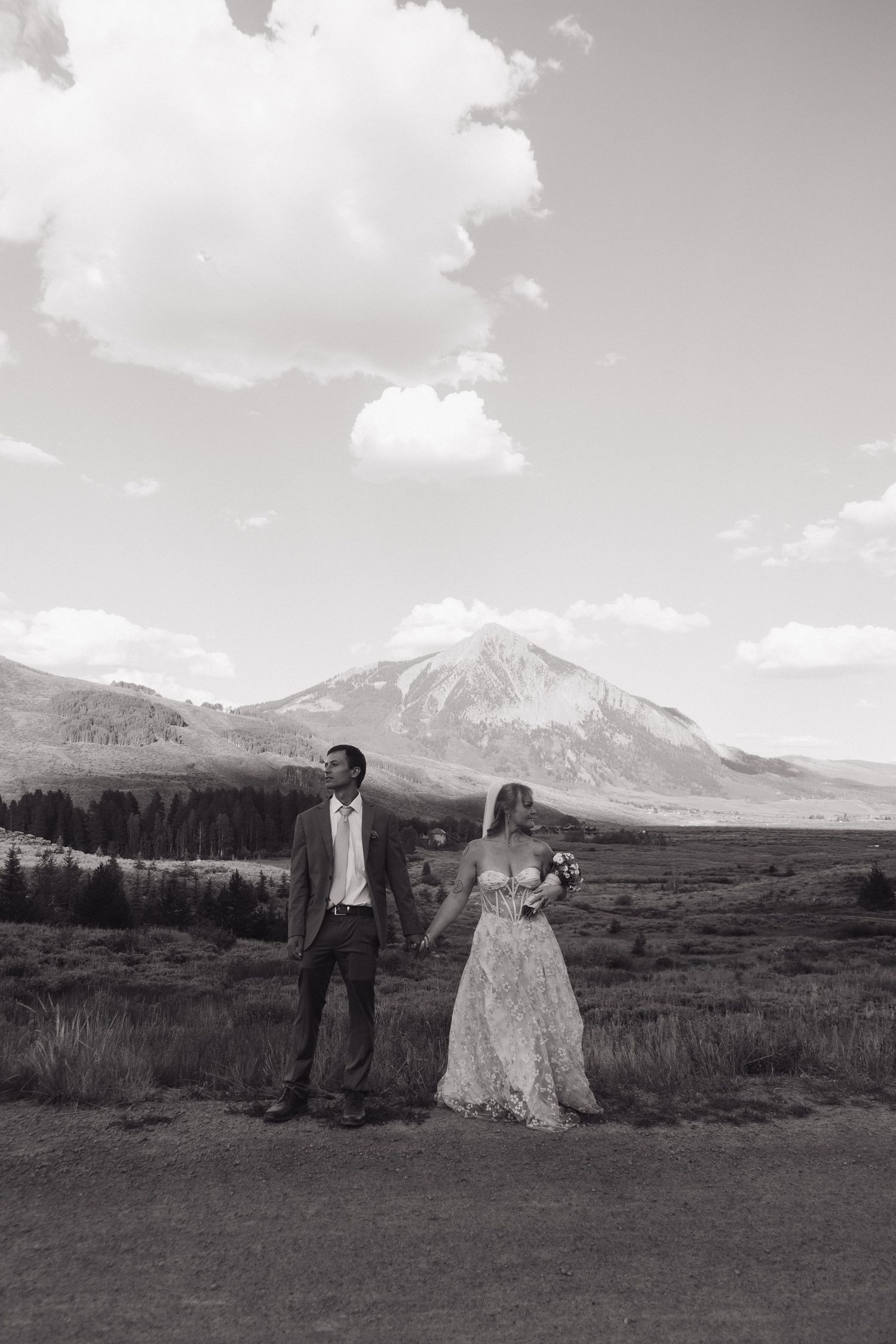 Black and white photo of bride and groom standing in mountain meadow with dramatic peak behind them.