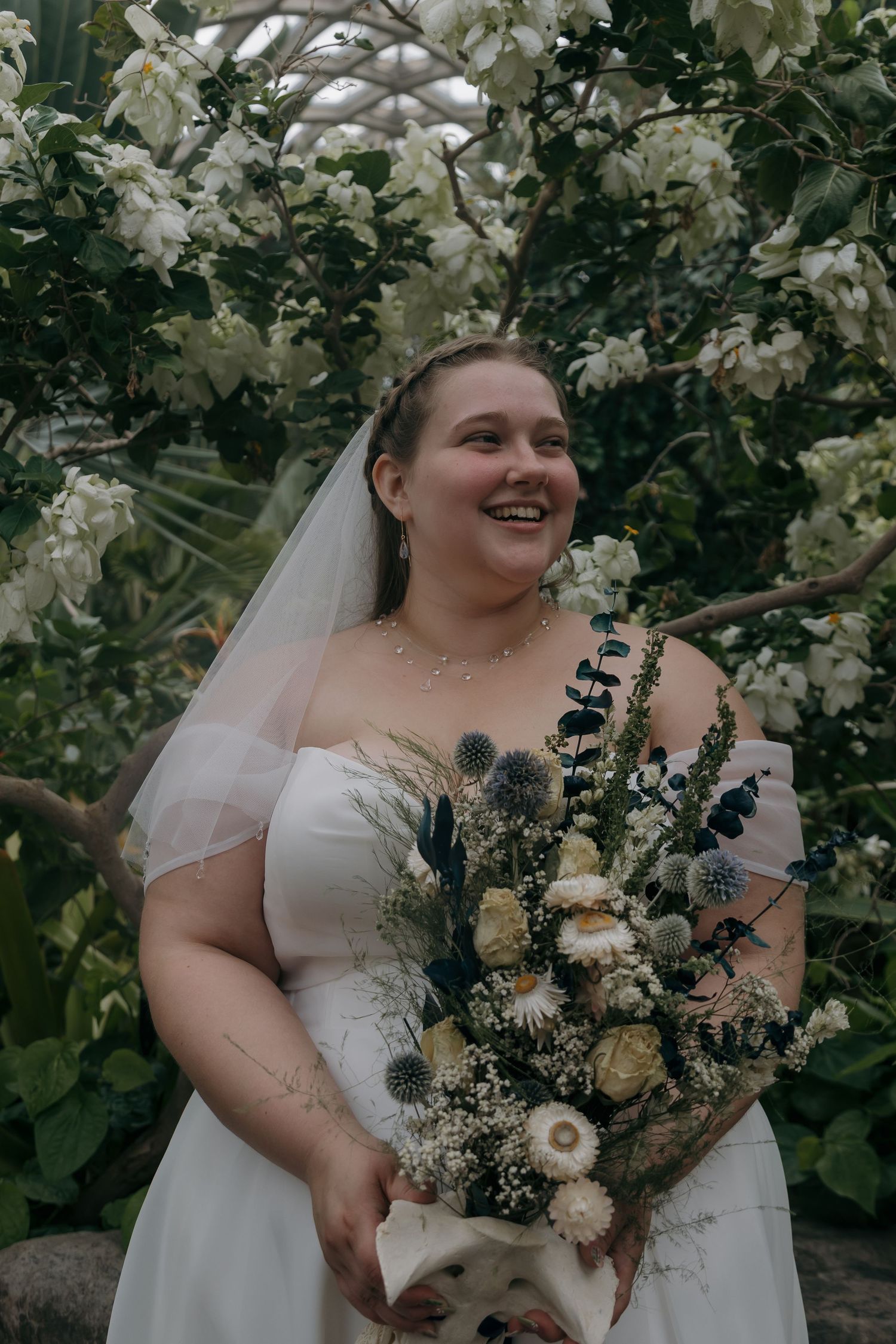 Smiling bride in white strapless gown and veil holds wildflower bouquet among lush white blooming trees.
