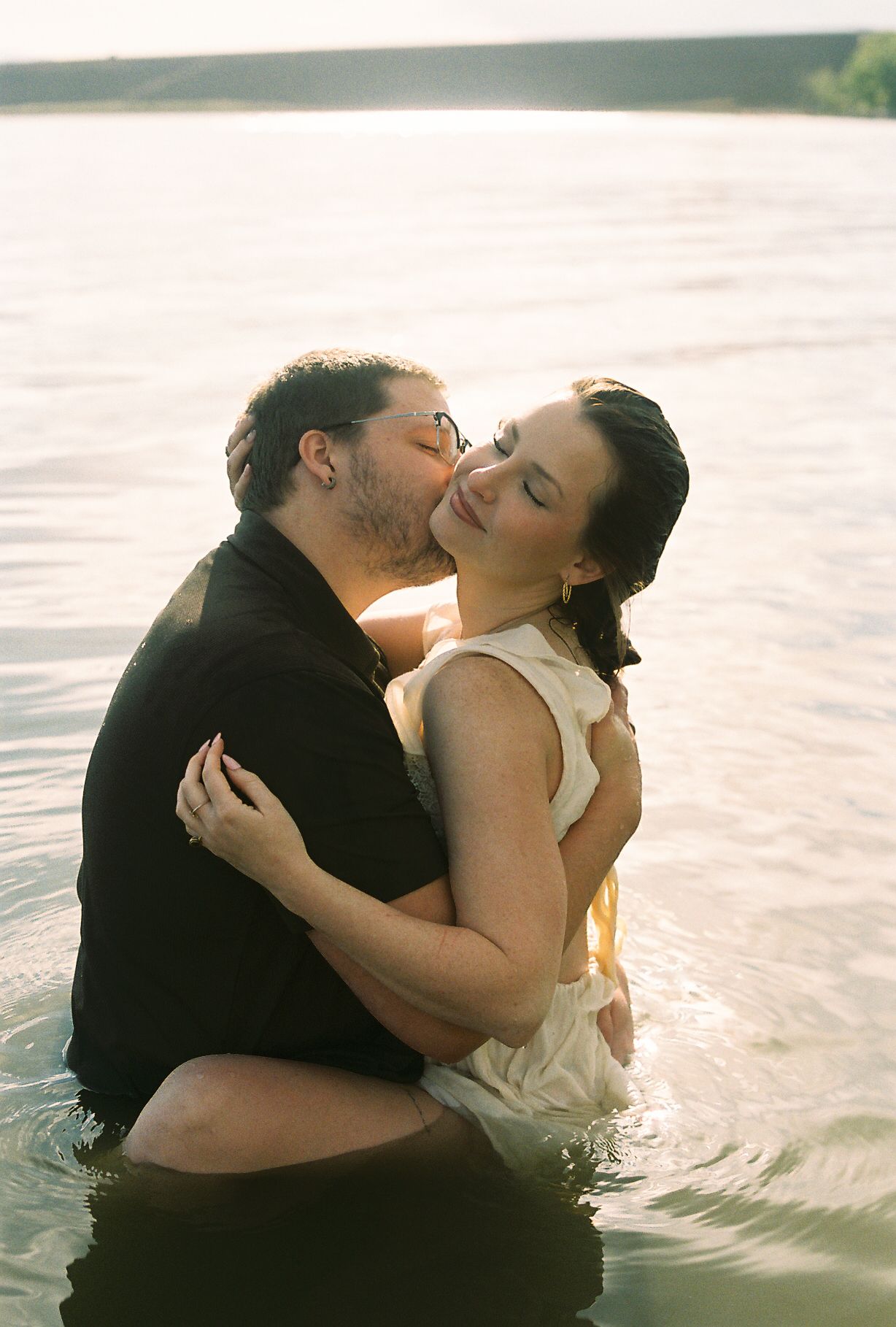 Couple sharing a romantic kiss while embracing in waist-deep lake water during a golden sunset session.