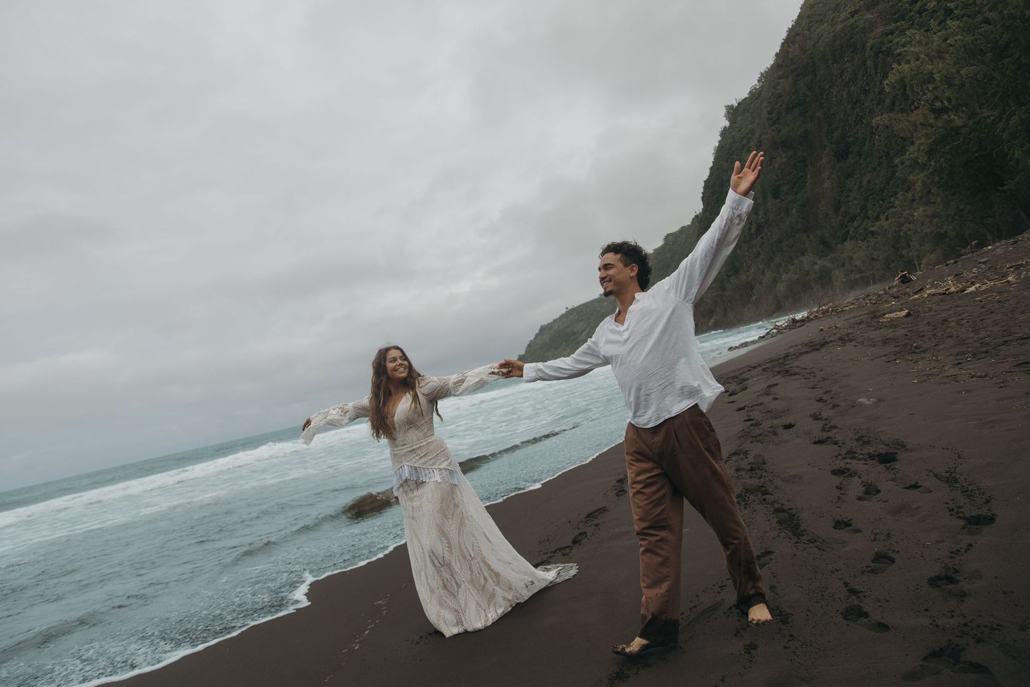 Couple in wedding attire dancing joyfully on a black sand beach with ocean waves and green cliffs behind them.
