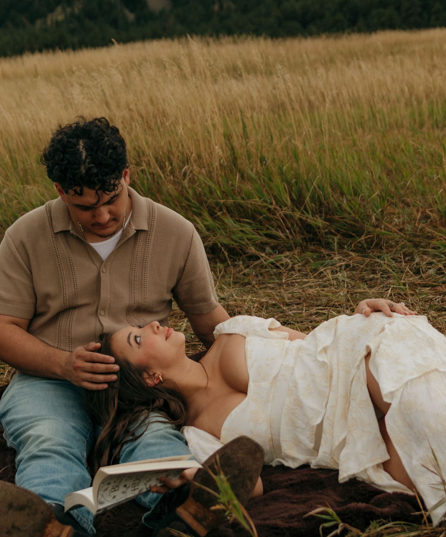 Romantic couple relaxing in golden grass field, woman lying down reading book with mountain backdrop.