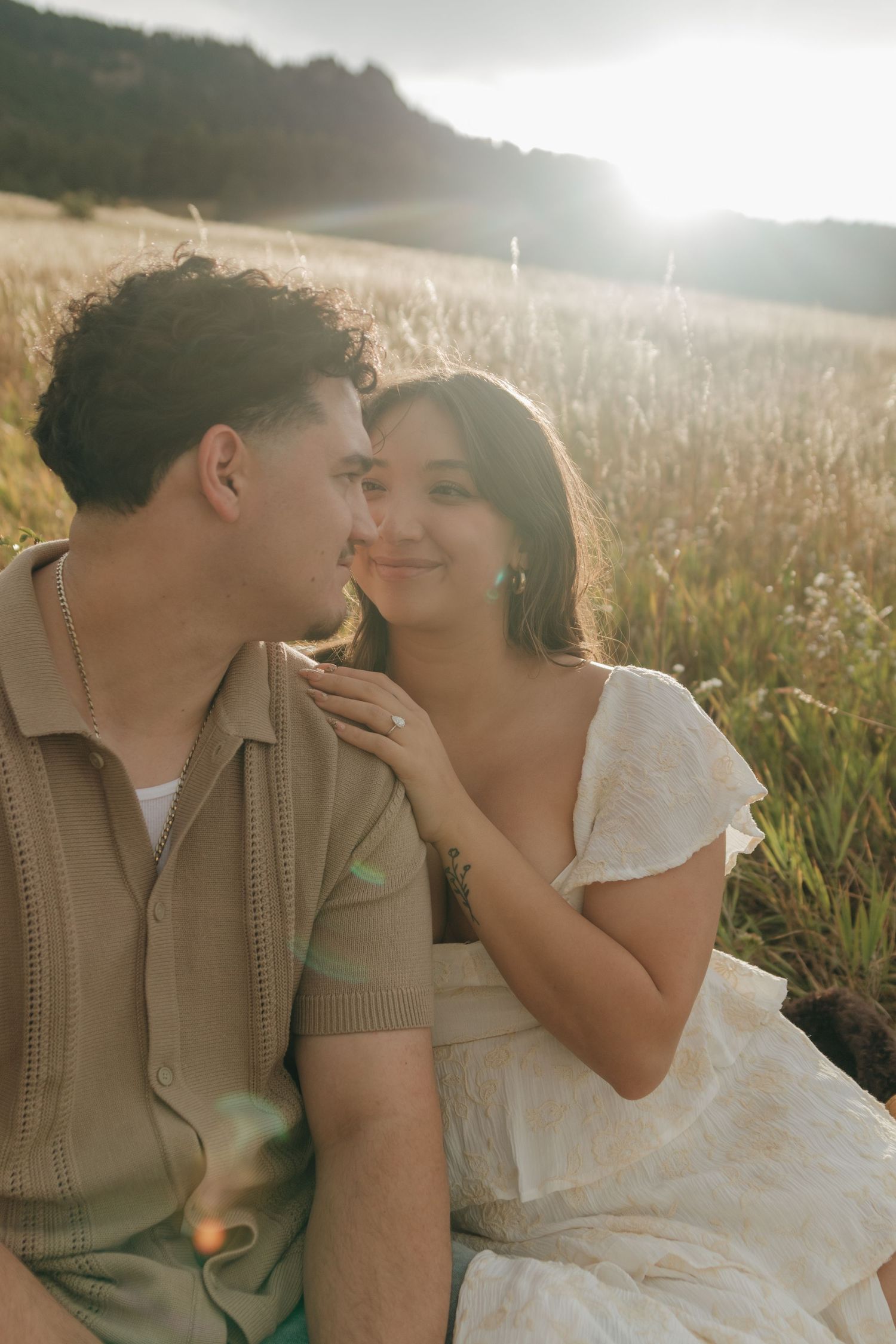 Couple sitting in golden field at sunset, woman in white dress smiling at man in beige shirt.