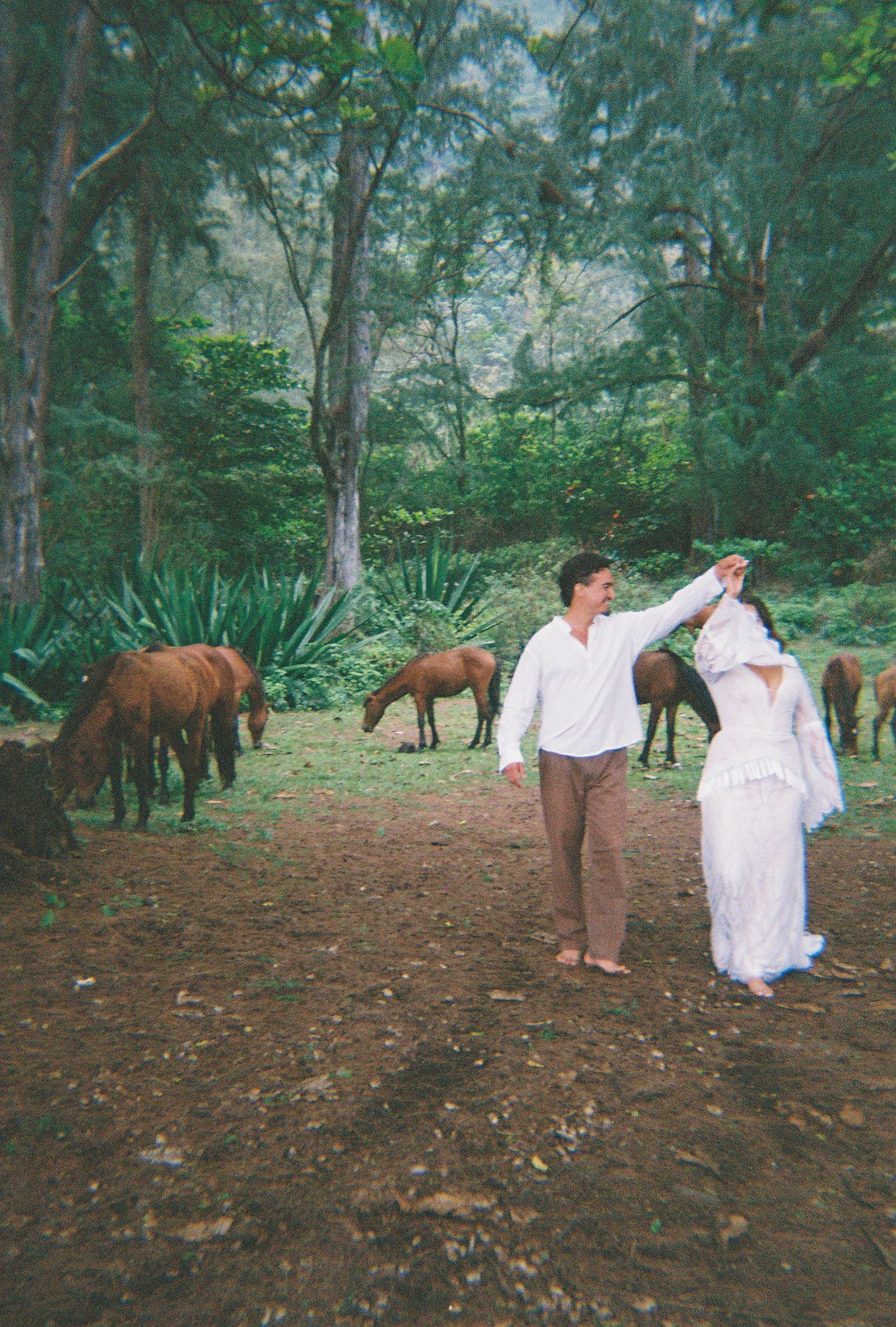 Couple dances among grazing horses in a lush green forest clearing in a film photo.