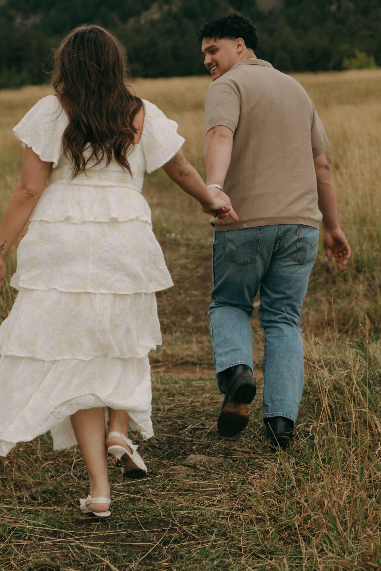 Couple holding hands walking through a golden meadow, woman in white ruffled dress.