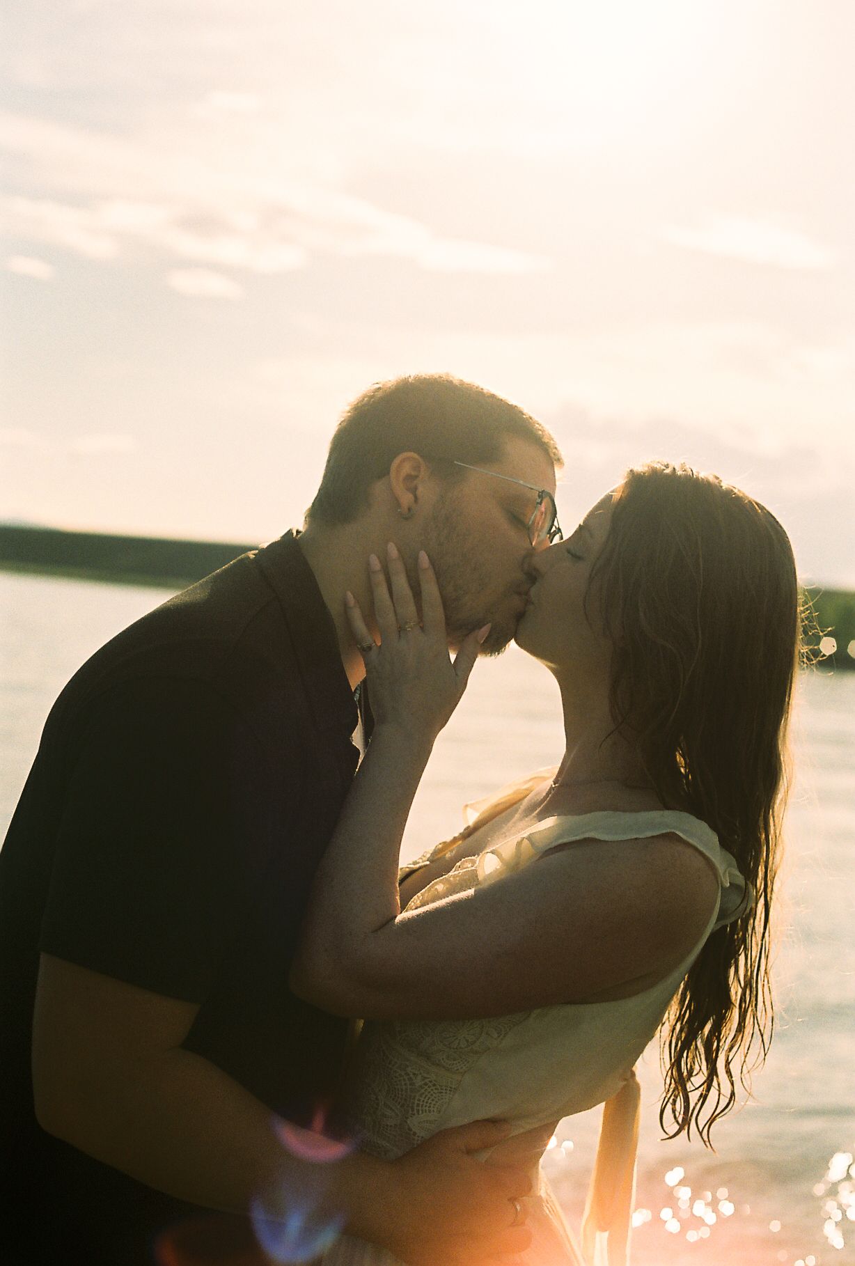 Couple sharing a passionate kiss backlit by a glowing sunset over the lake.
