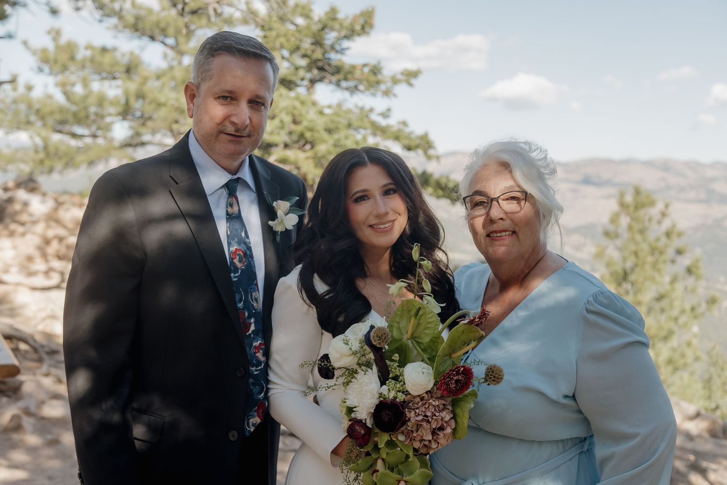 Bride posing with parents at outdoor mountain wedding surrounded by pine trees and scenic landscape.