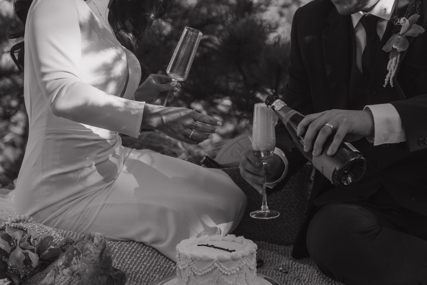 Black and white photo of a couple pouring champagne at an outdoor wedding picnic celebration.