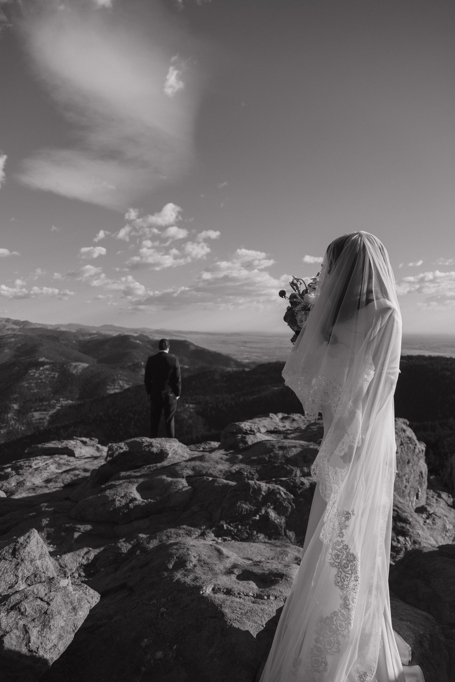 Black and white photo of bride holding bouquet watching groom gaze over mountain landscape.