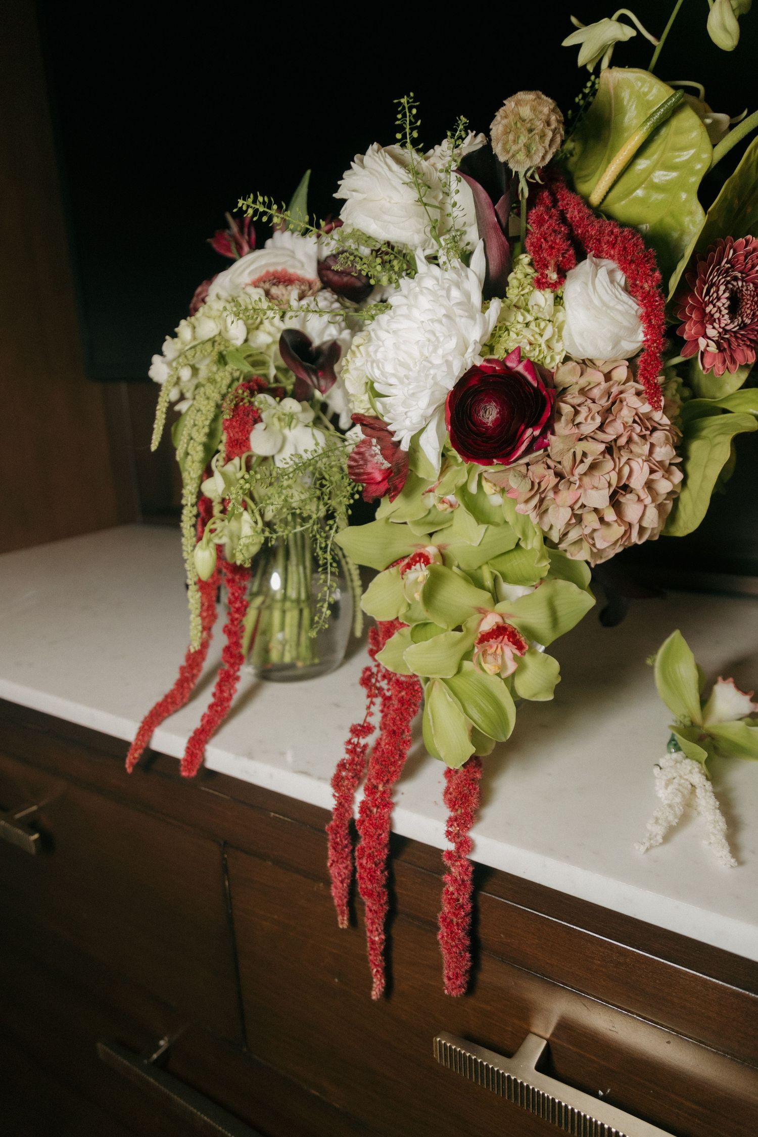 Lush wedding bouquet featuring green orchids, white chrysanthemums, red amaranthus, and dusty pink hydrangeas on a marble surface.