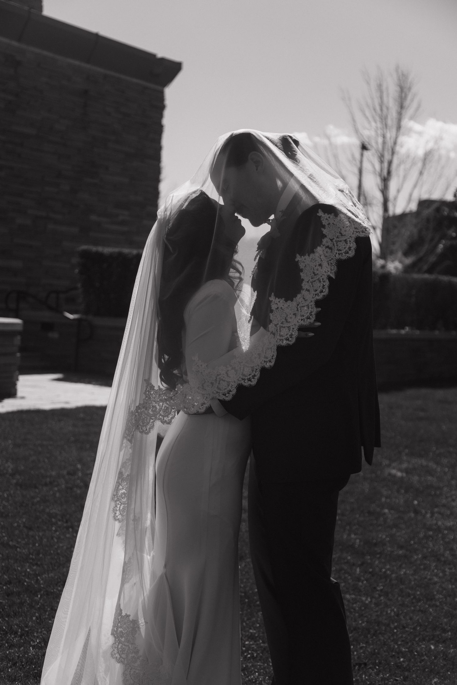 Black and white photo of bride and groom embracing under lace-trimmed veil outdoors.