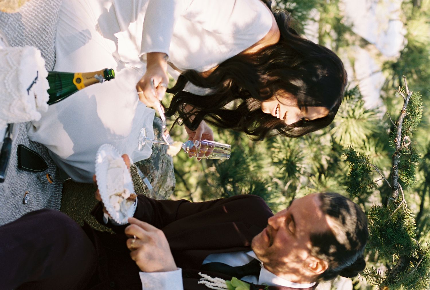 Bride and groom enjoy champagne and wedding cake together outdoors among pine trees after ceremony.