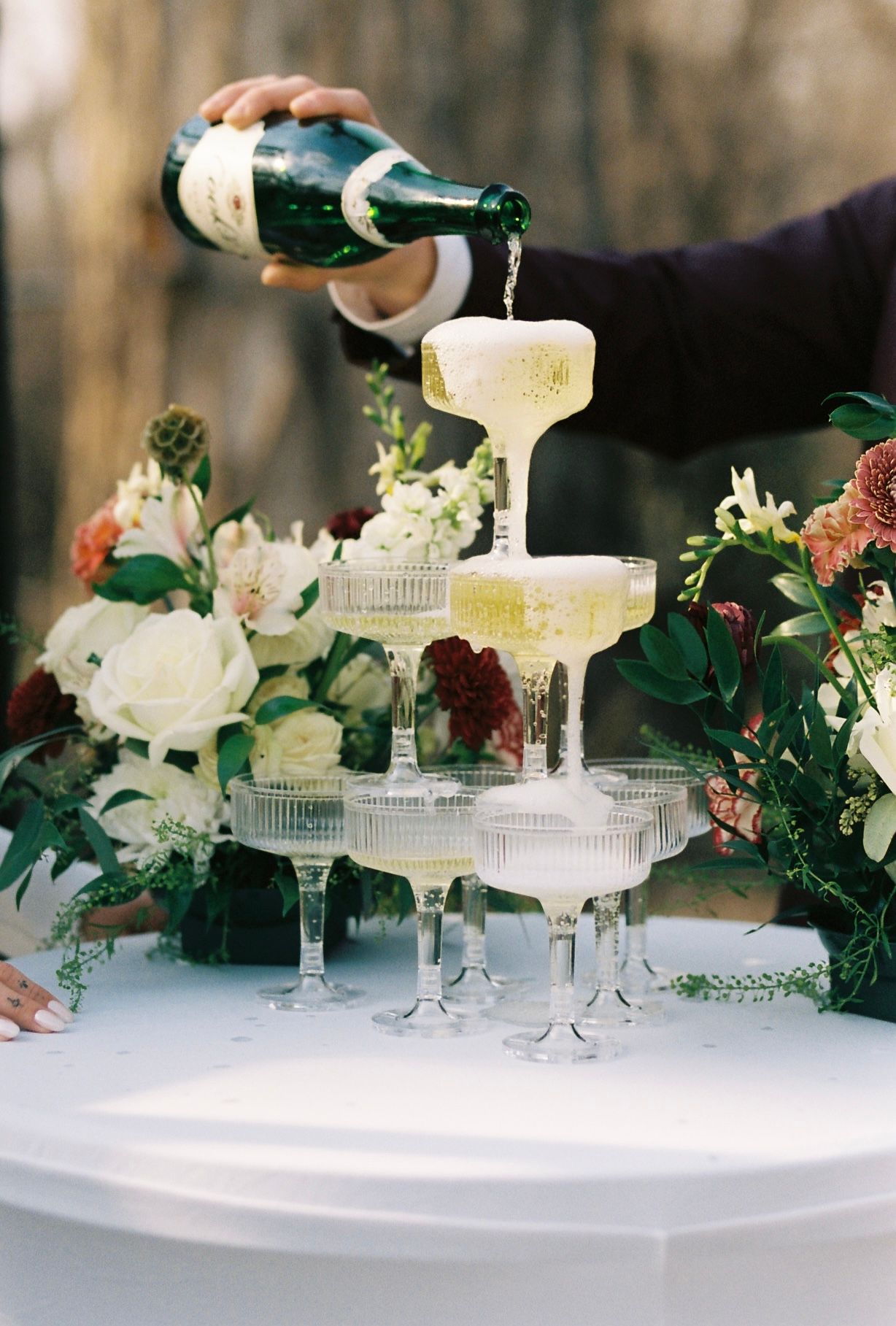 Champagne tower being poured at an elegant outdoor wedding reception with floral arrangements.