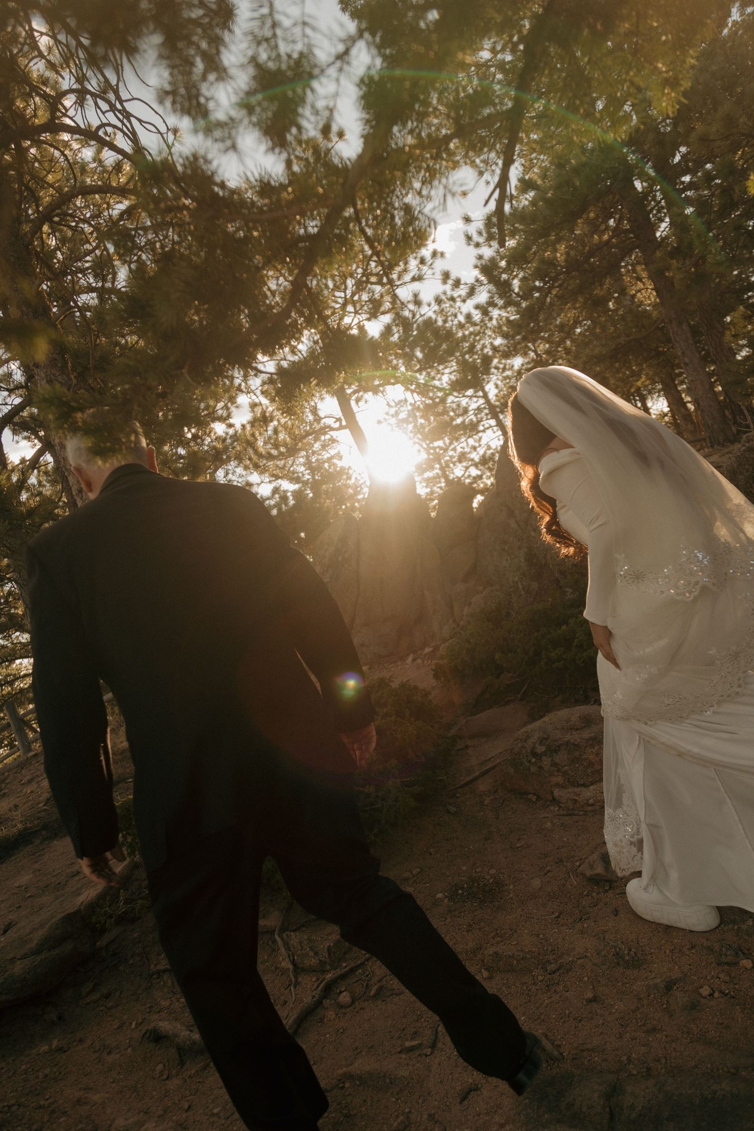 Bride in white gown and groom walk through sunlit pine forest during elopement.