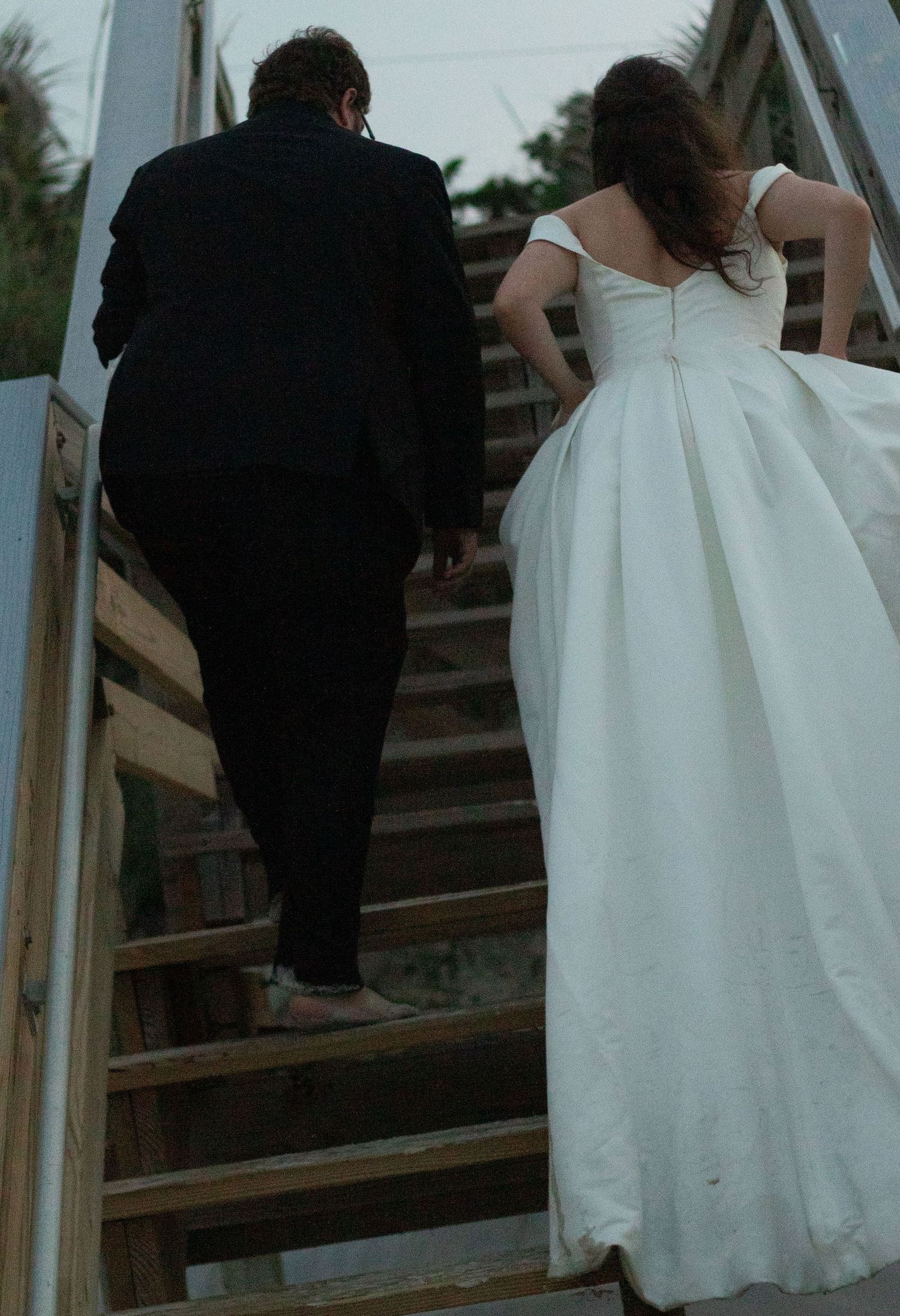 A couple in wedding attire climbs wooden stairs outdoors during a rustic ceremony.