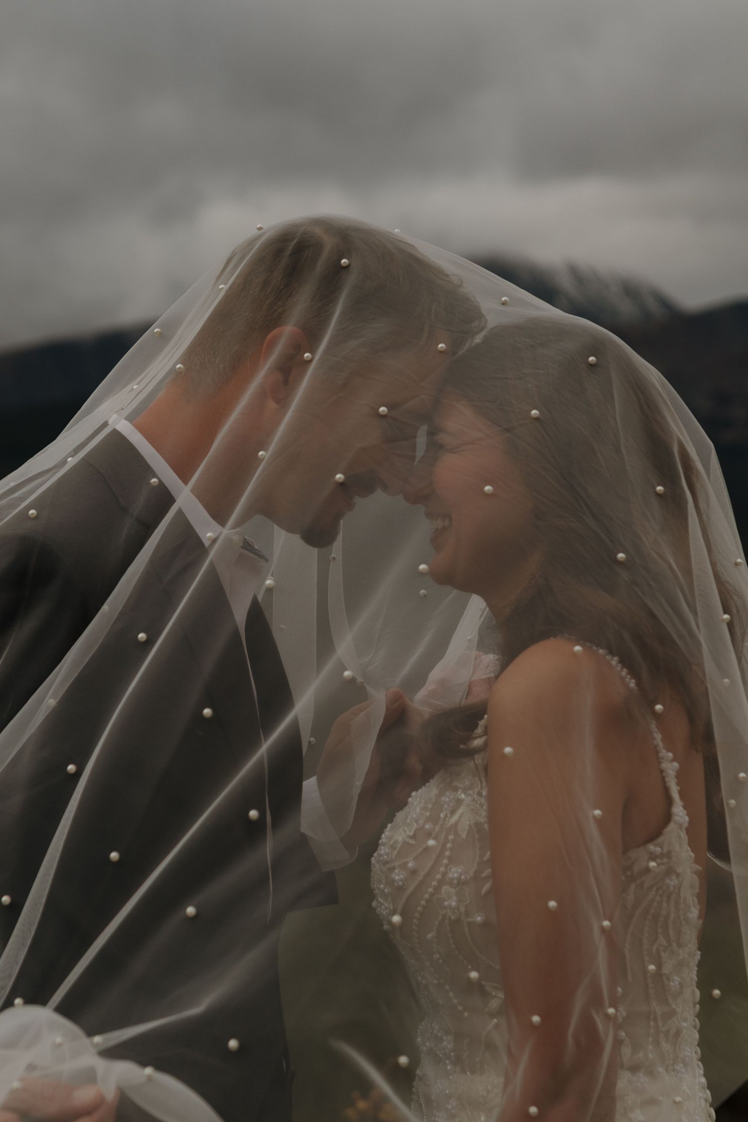 Newlyweds share a tender moment beneath a flowing veil in a dramatic outdoor setting.