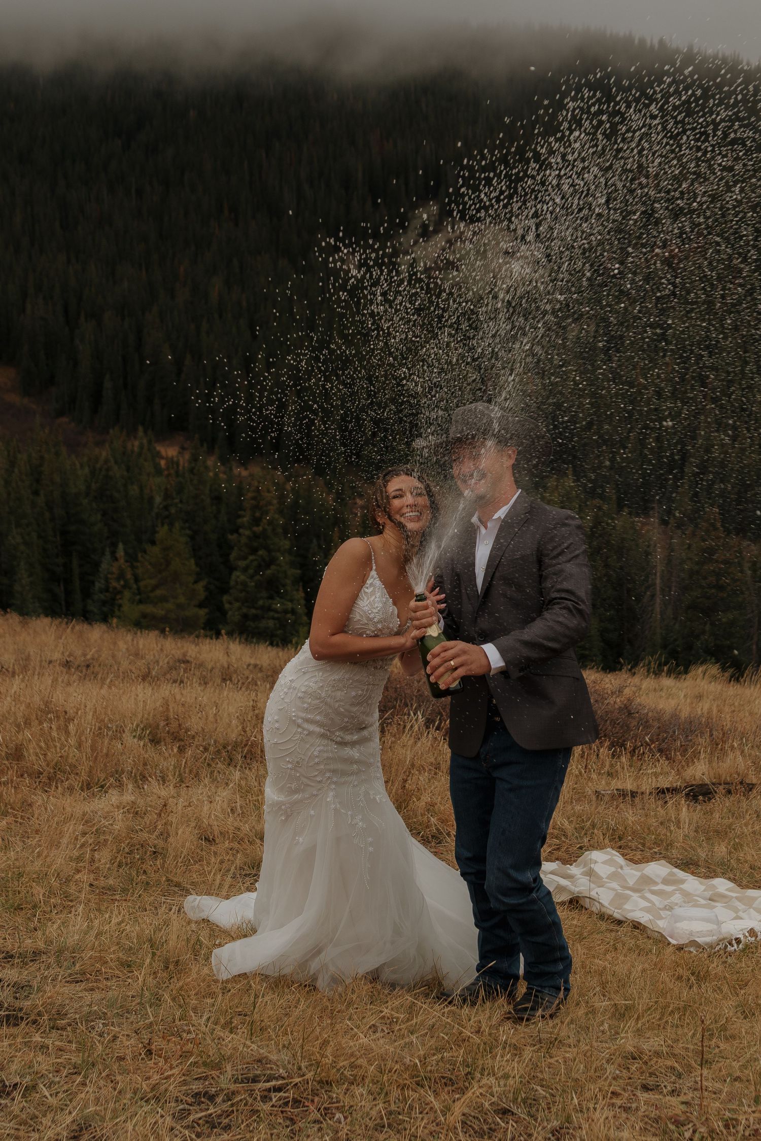 Newlyweds pop champagne on a mountainside during their rustic outdoor wedding celebration.