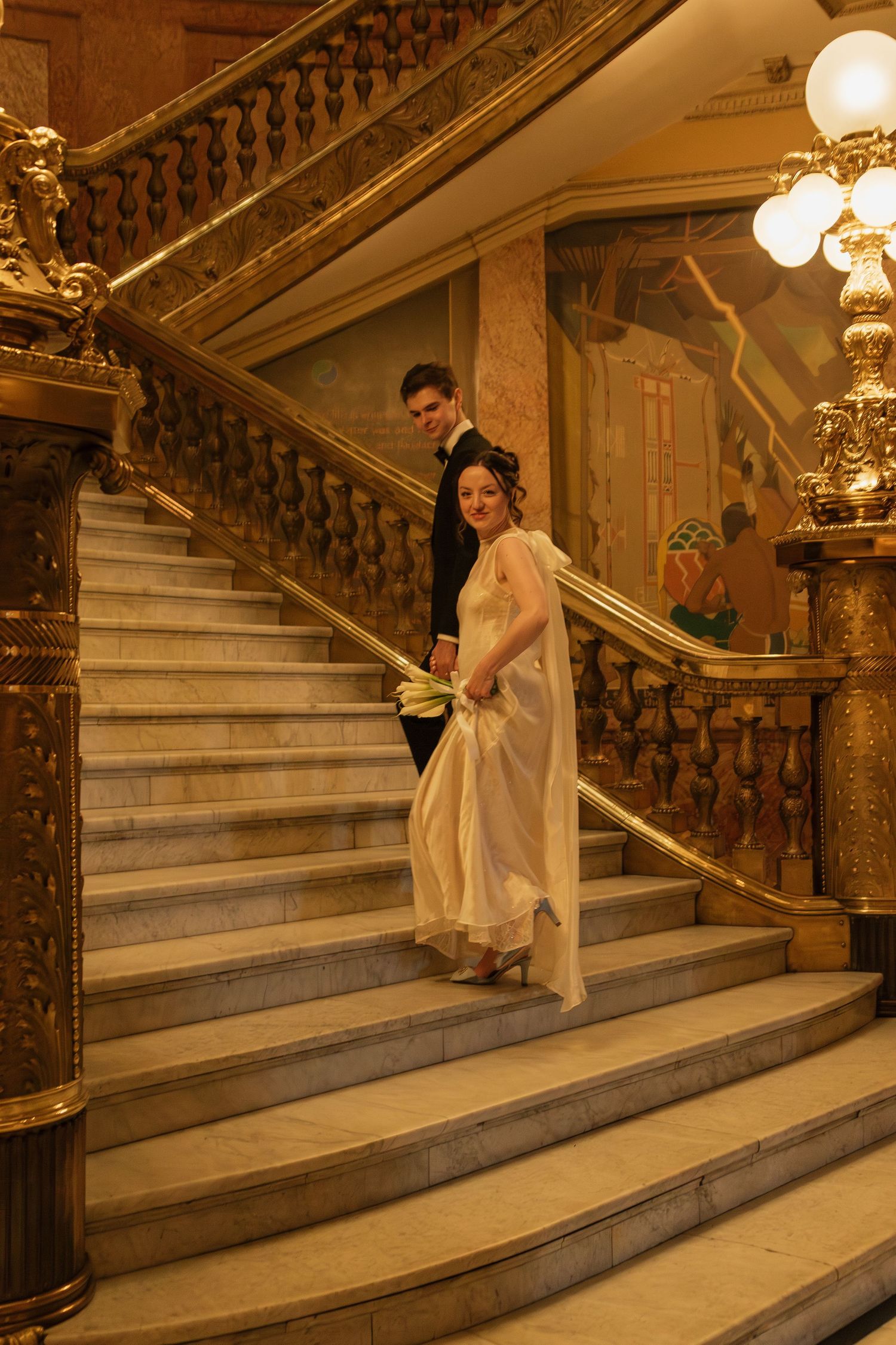 Couple posing on grand marble staircase with ornate railings and vintage lighting fixtures.