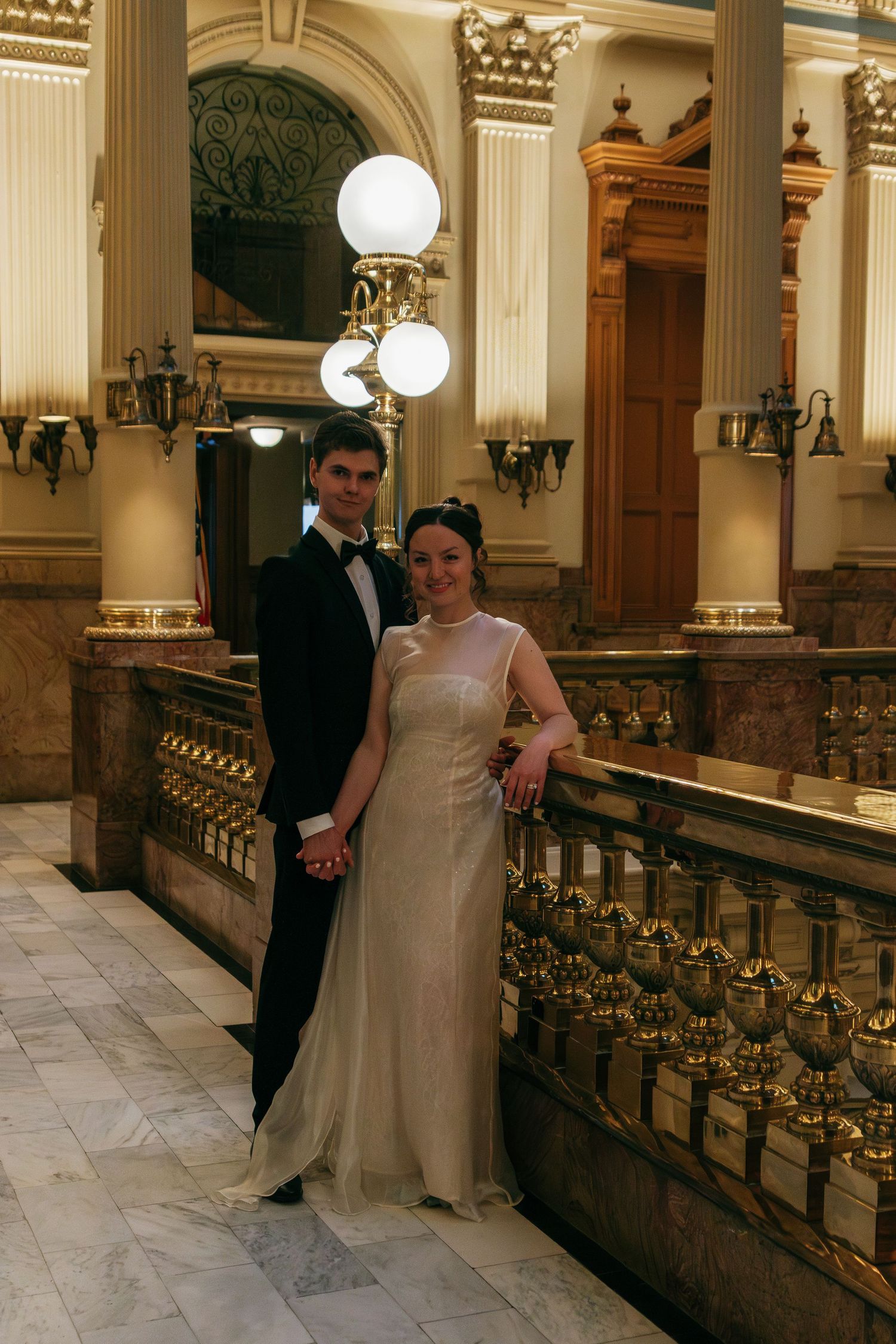 A couple poses together in a grand marble hallway with ornate railings and classical architecture.