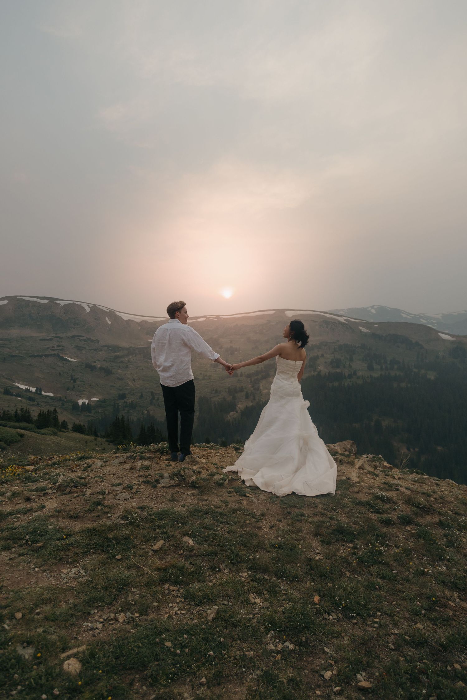 Bride and groom holding hands on mountain summit at sunset, overlooking scenic valley landscape.