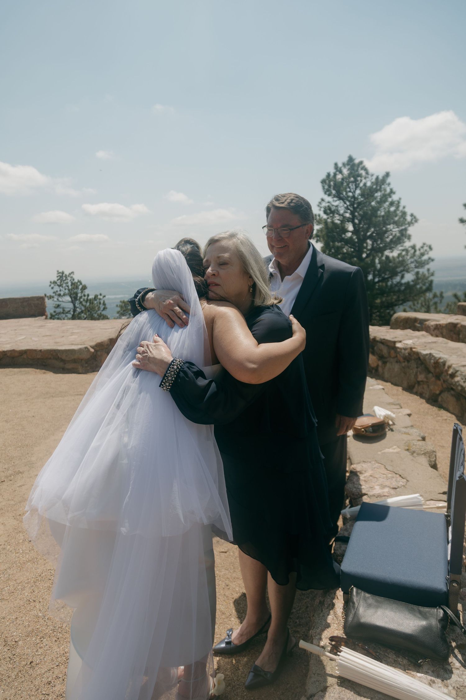 A bride in a white veil shares an emotional hug with a woman while a man in a dark suit stands nearby outdoors.