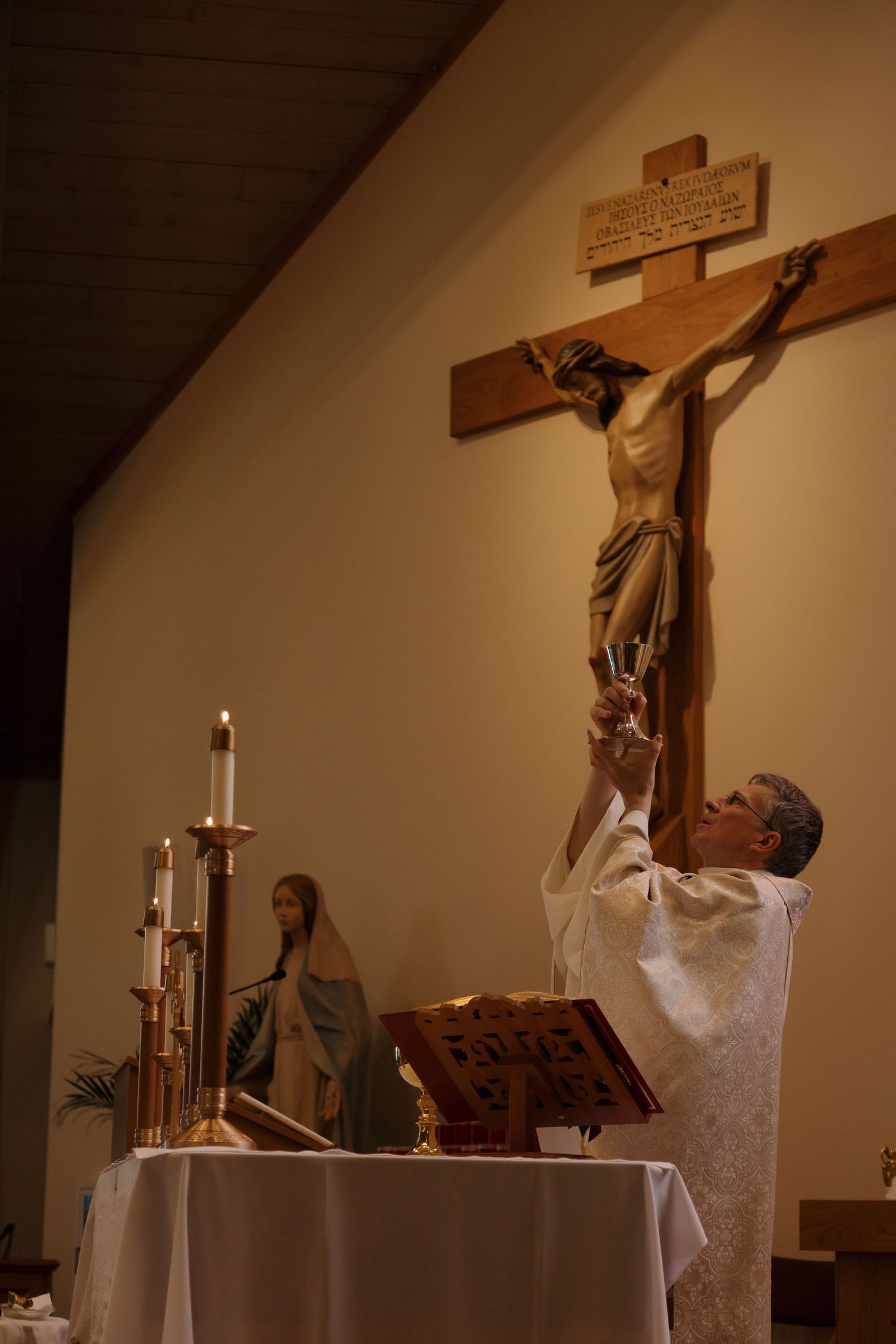 A priest elevates the chalice before a crucifix during a candlelit Catholic Mass ceremony.