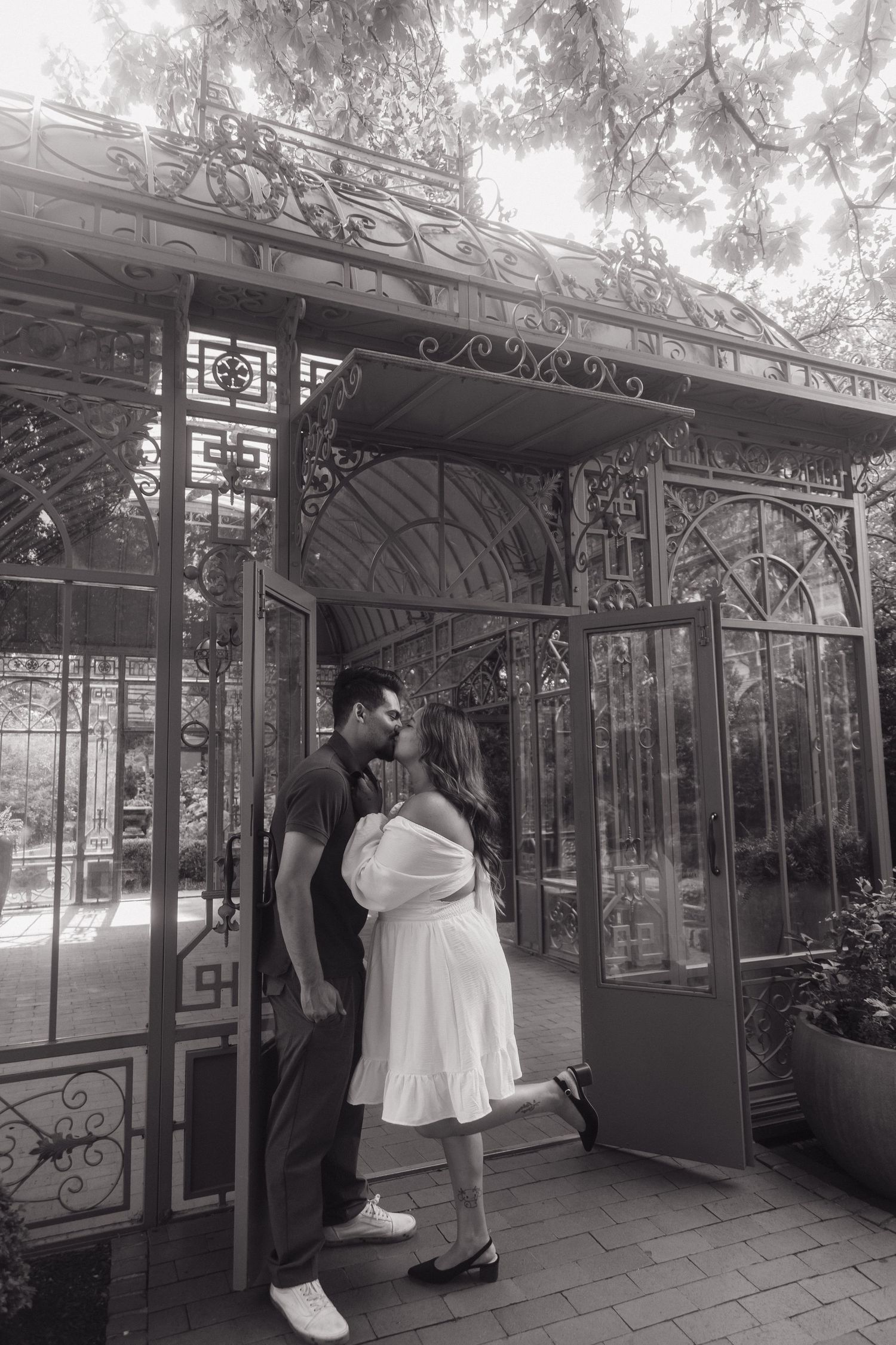 Black and white photo of couple kissing at the entrance of an ornate iron greenhouse structure.