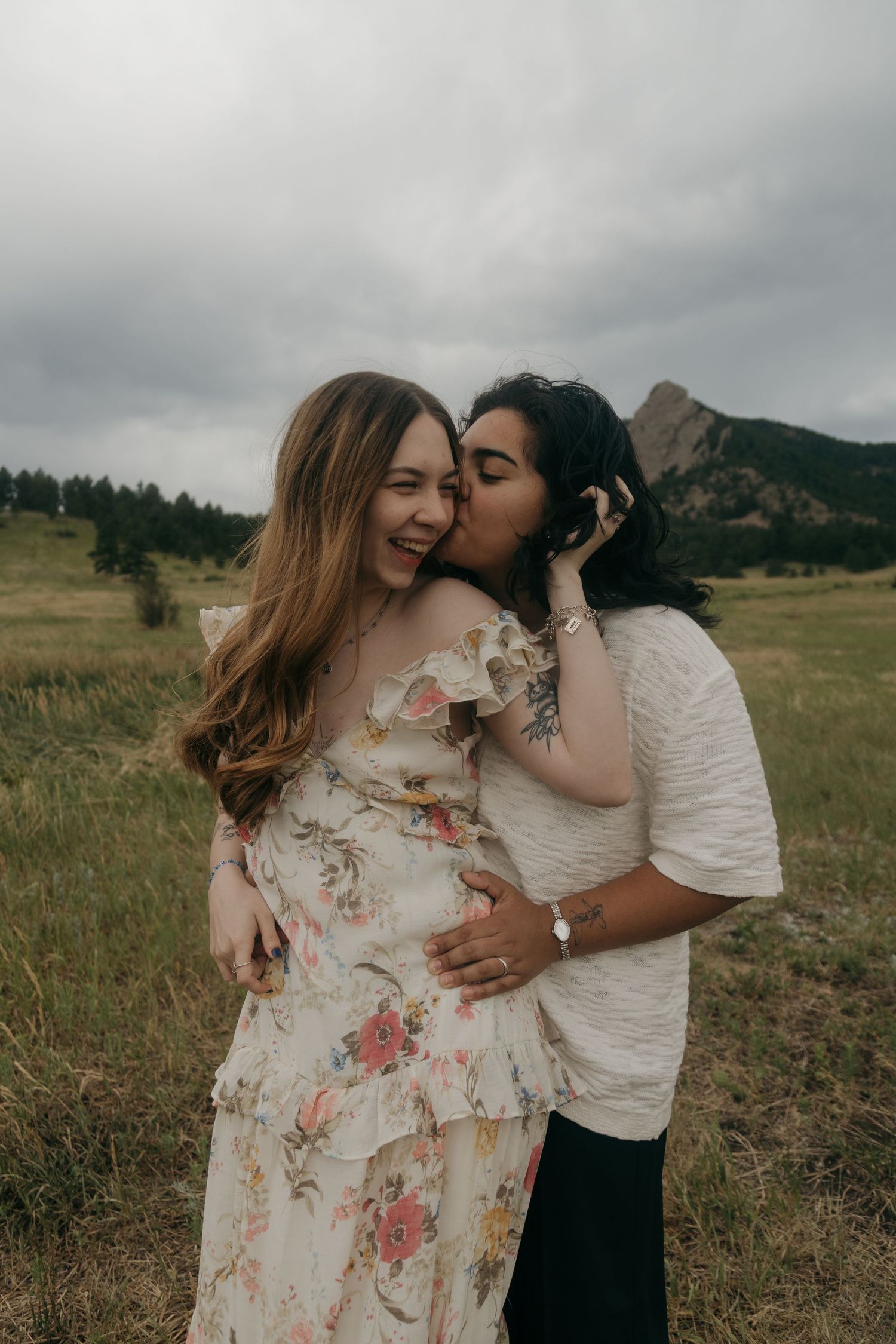 A woman in a floral dress laughs as her partner kisses her cheek in a scenic mountain meadow.