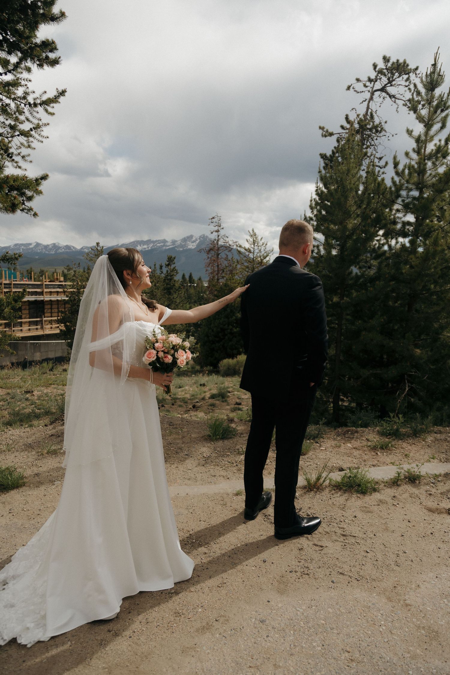 Bride in white gown and veil taps groom shoulder for first look with mountain views.