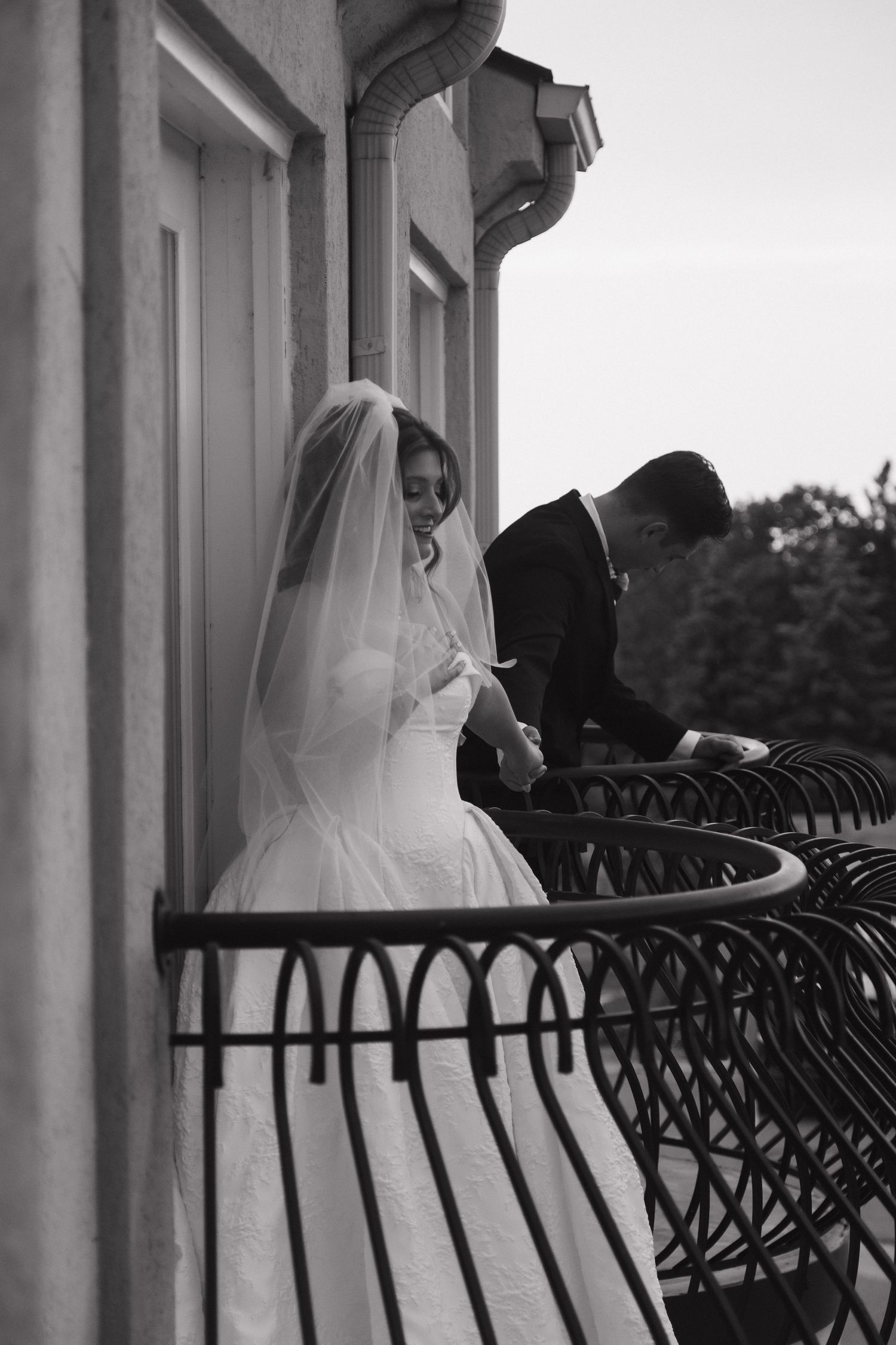 Black and white photo of a bride in veil and groom on an ornate balcony on their wedding day.