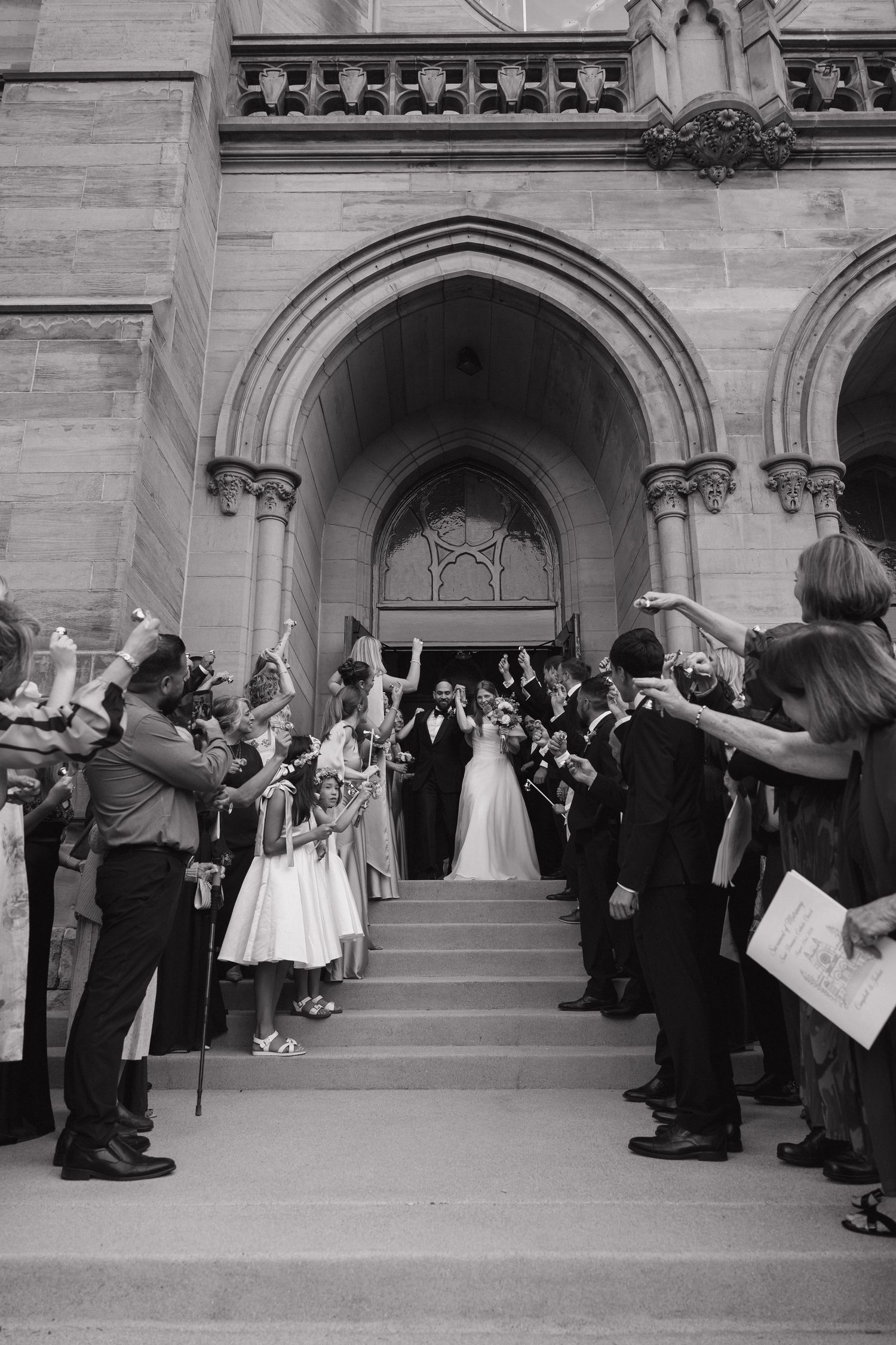 Newlyweds exit grand stone church greeted by cheering guests in black and white photo.