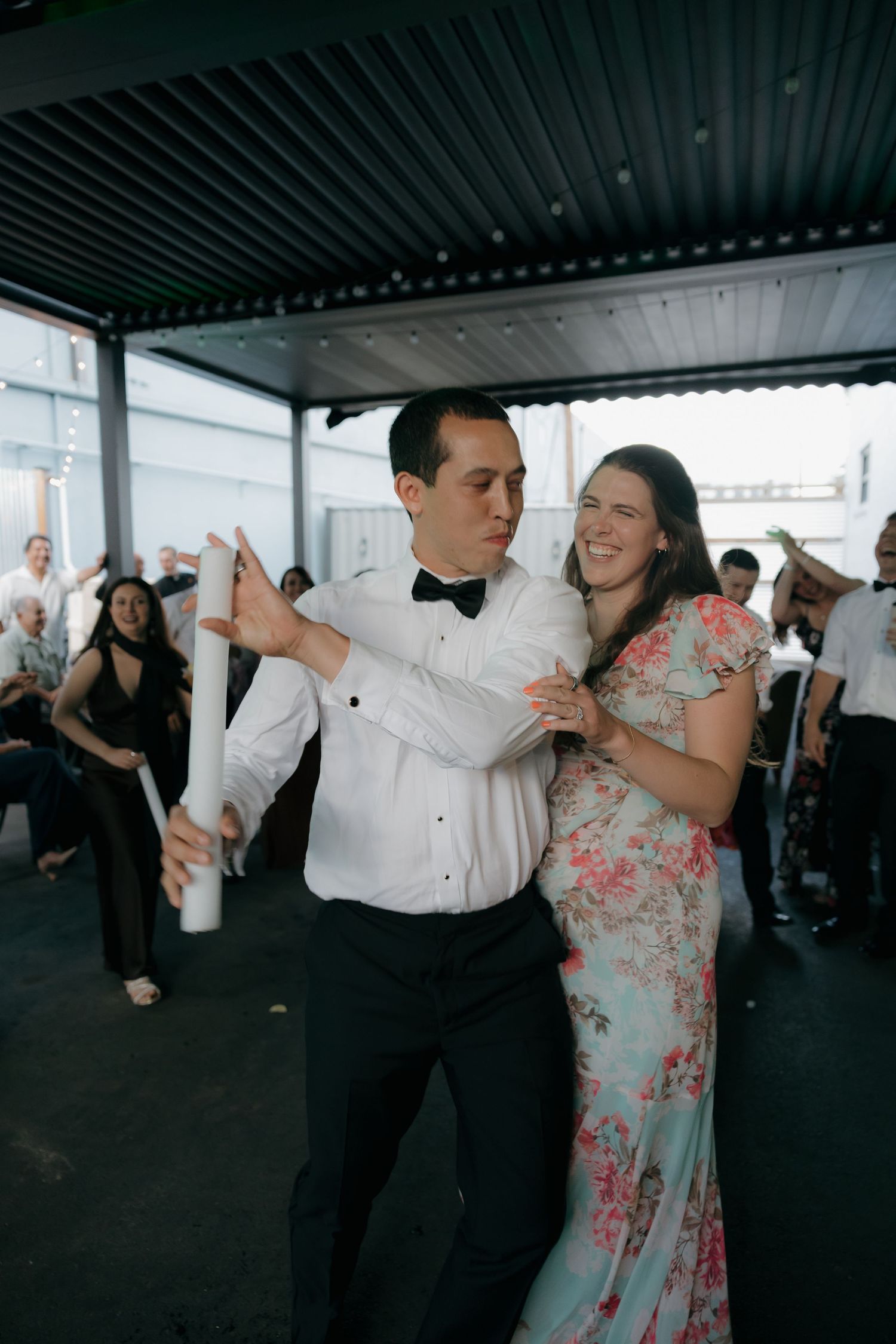 Groom in bow tie dances with smiling woman in floral dress while guests celebrate at outdoor wedding reception.