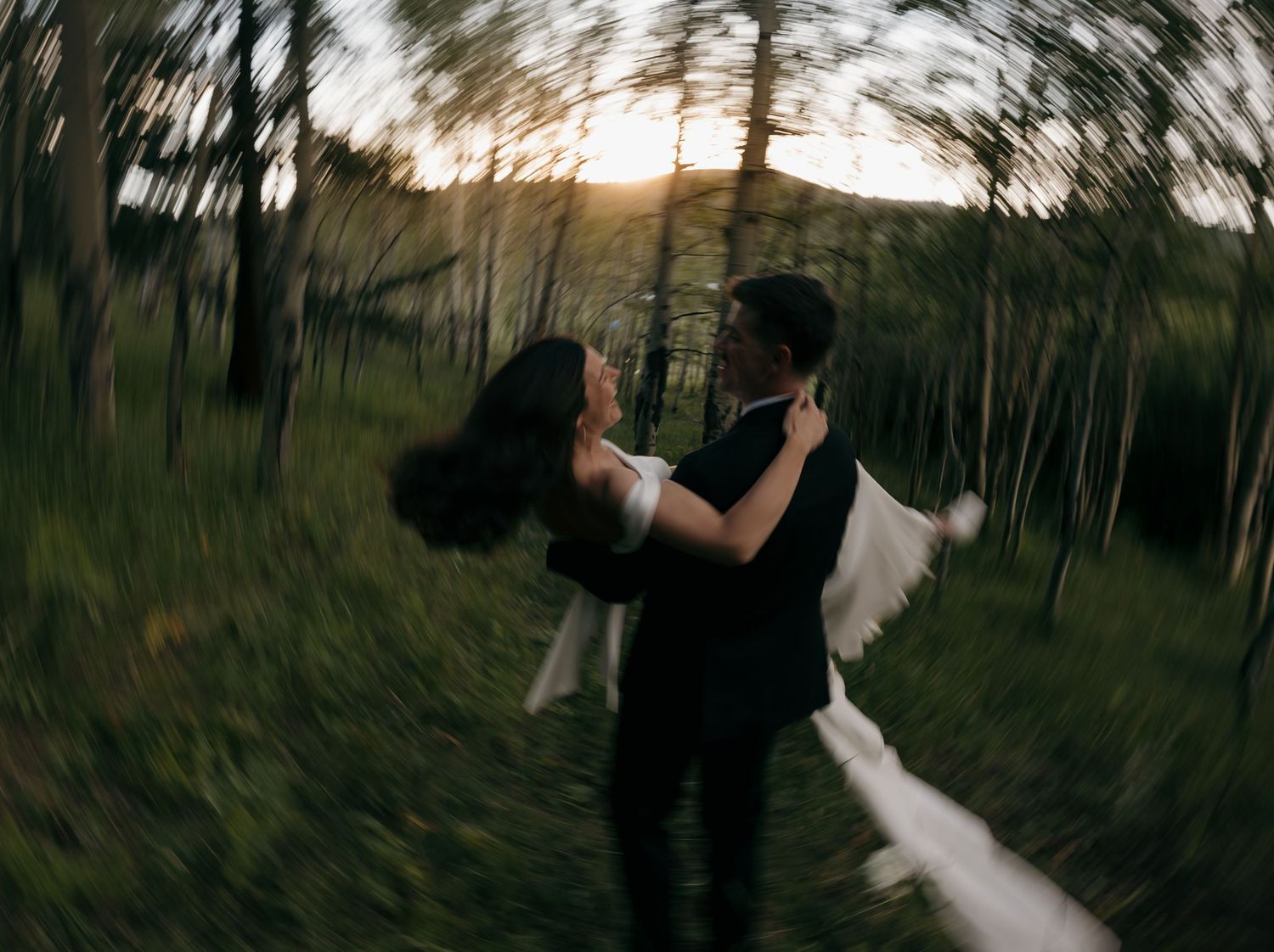 Groom lifting bride in a sunlit forest, captured with a swirling motion blur effect at golden hour.