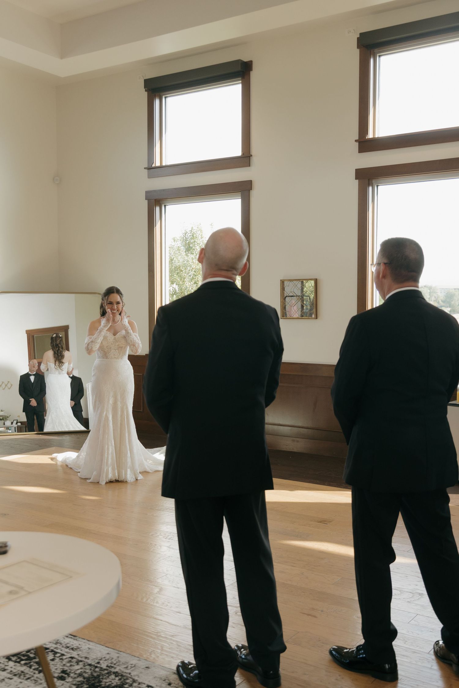 Bride in white gown shares emotional first look moment with two men in black suits indoors.
