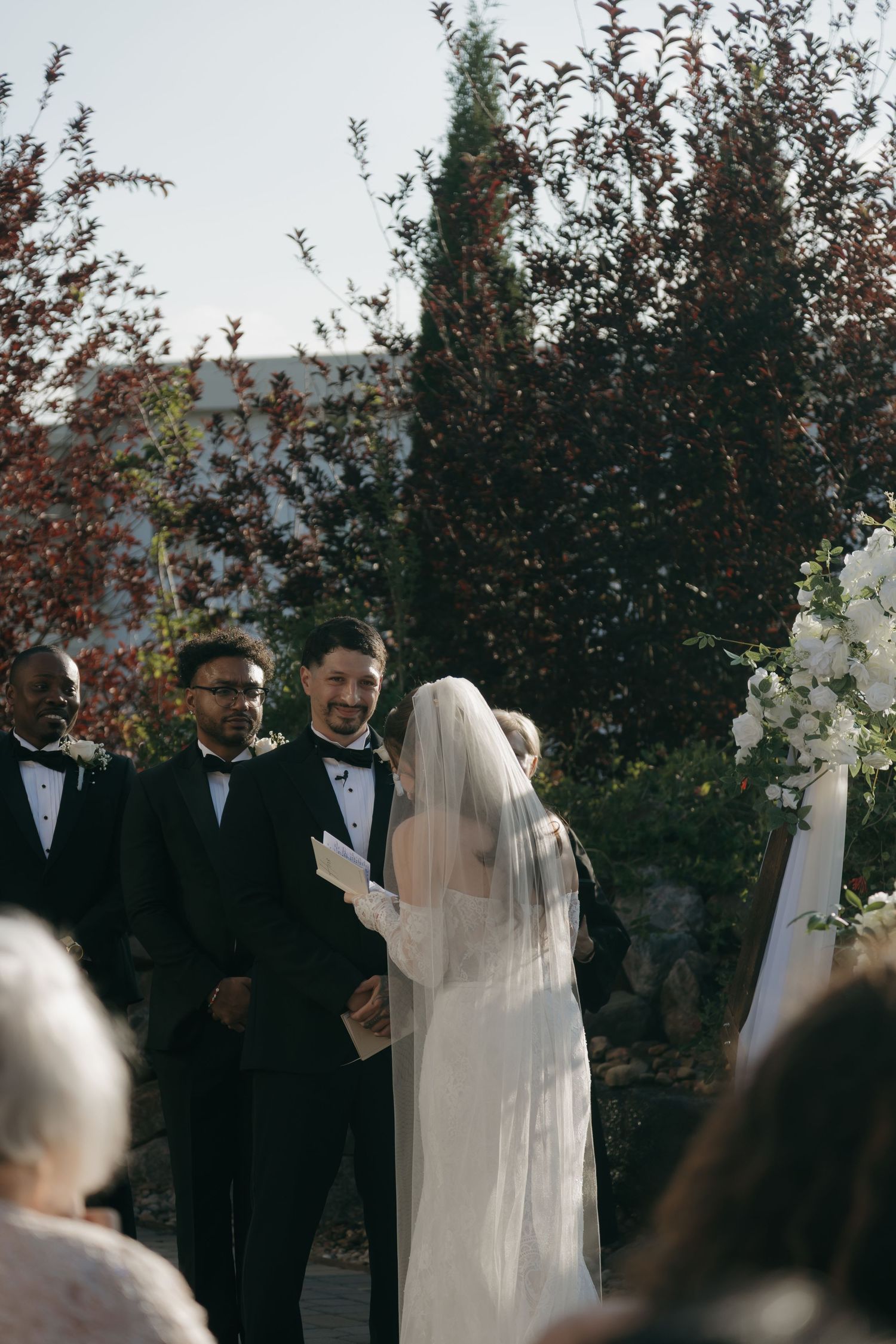 Groom smiling at veiled bride during outdoor wedding ceremony with white floral arch backdrop.